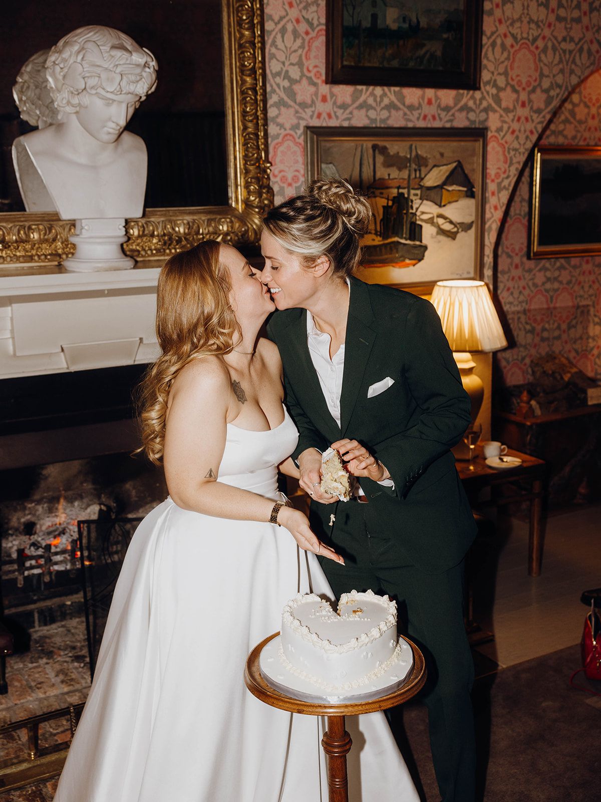 A couple shares an intimate moment while cutting their wedding cake in an elegant room with ornate decor and a marble bust.