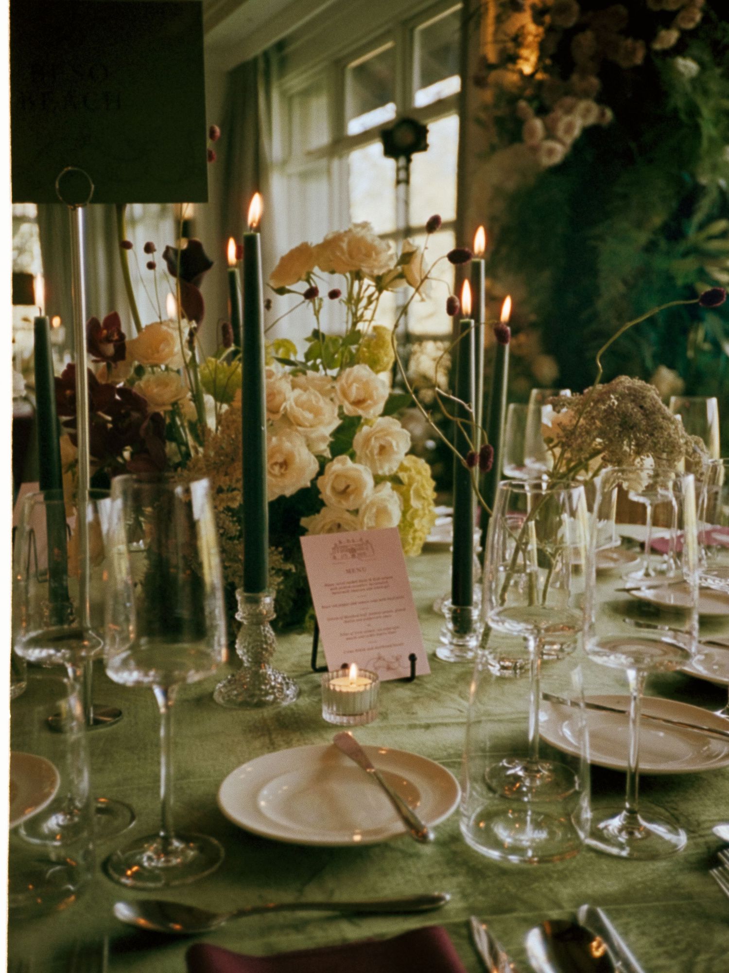 Elegant wedding table setting with green candles, white roses, crystal glassware, and a pink menu card on a green tablecloth.