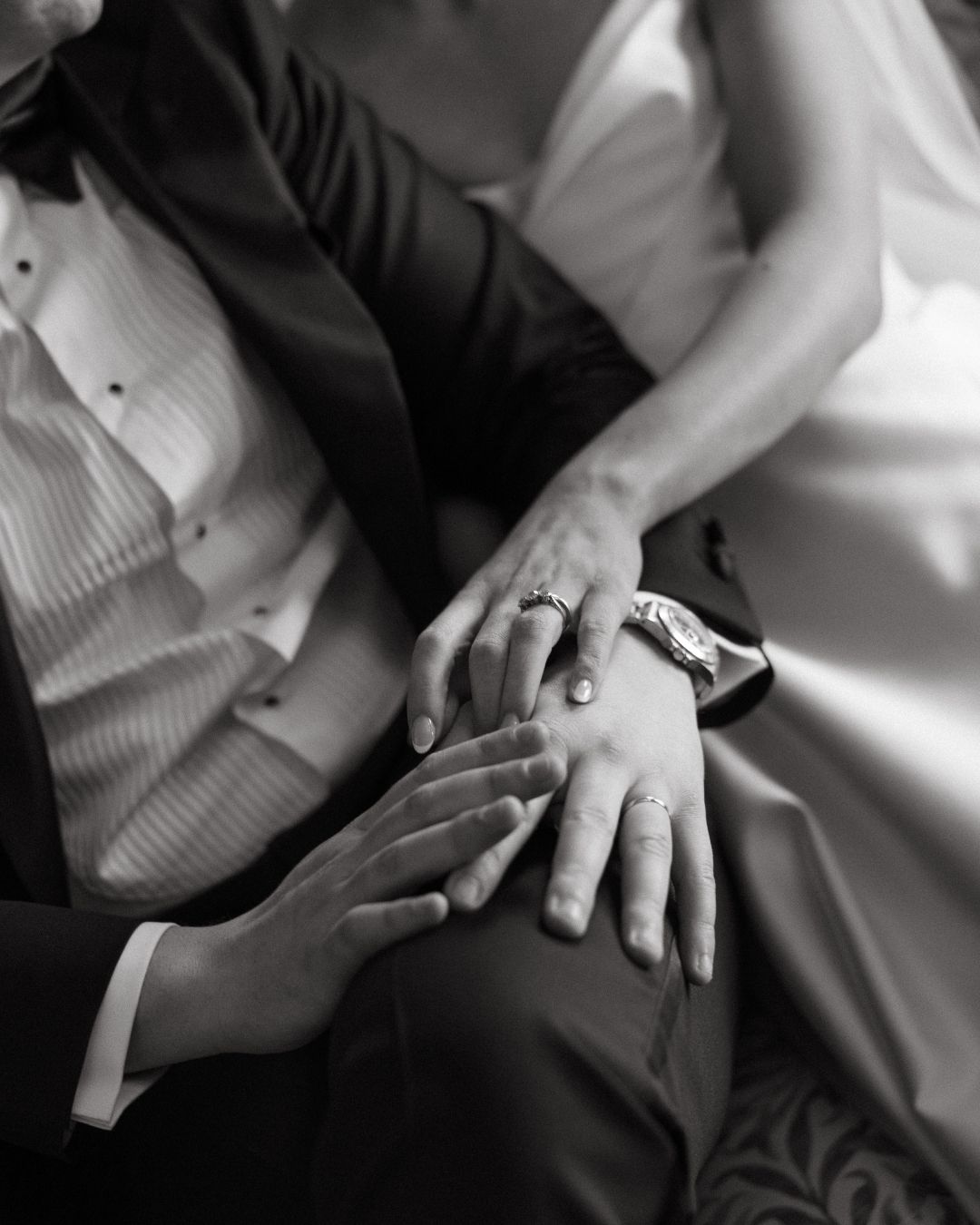 Black and white wedding photo of bride and groom hands showing rings and watch while seated together.
