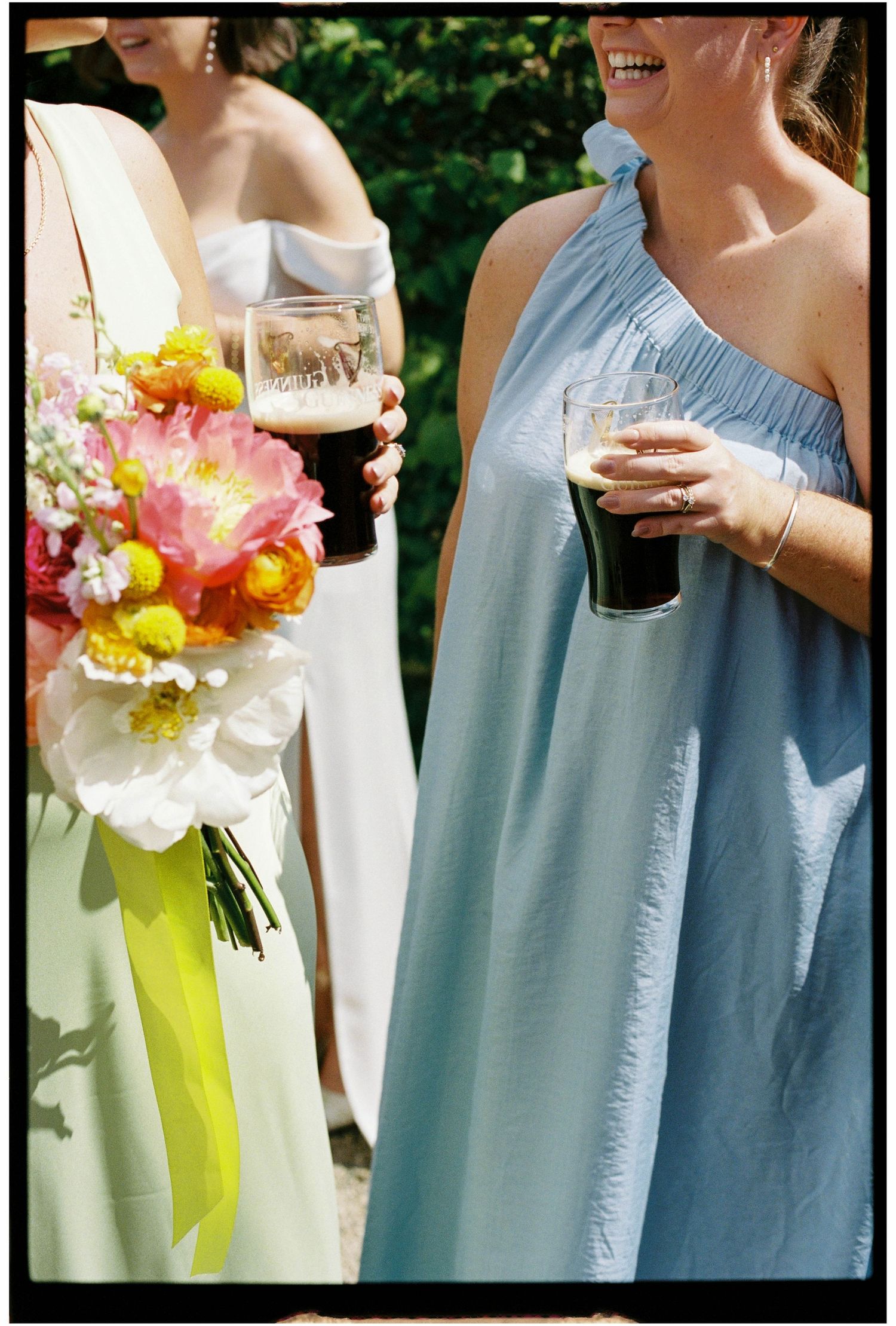 Bridesmaids in mint and blue dresses hold dark beer glasses alongside a vibrant pink and yellow wedding bouquet.