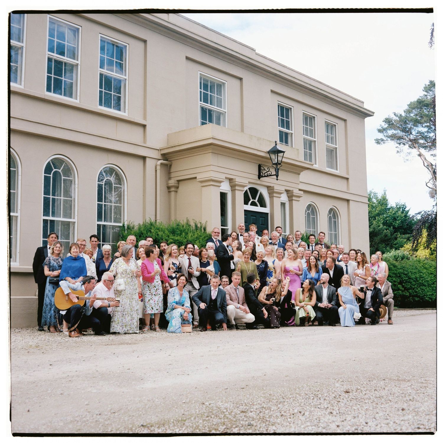 Large wedding group photo in front of an elegant Georgian manor house, guests dressed in colorful formal attire.