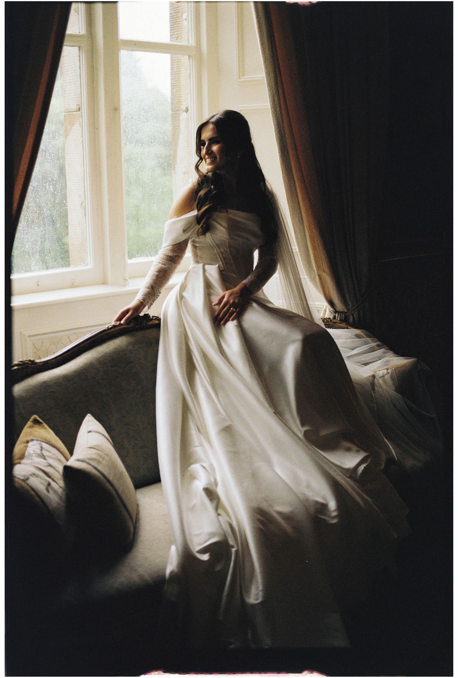 Bride in elegant off-shoulder white wedding gown standing by a sunlit window, with flowing skirt and long sleeves.