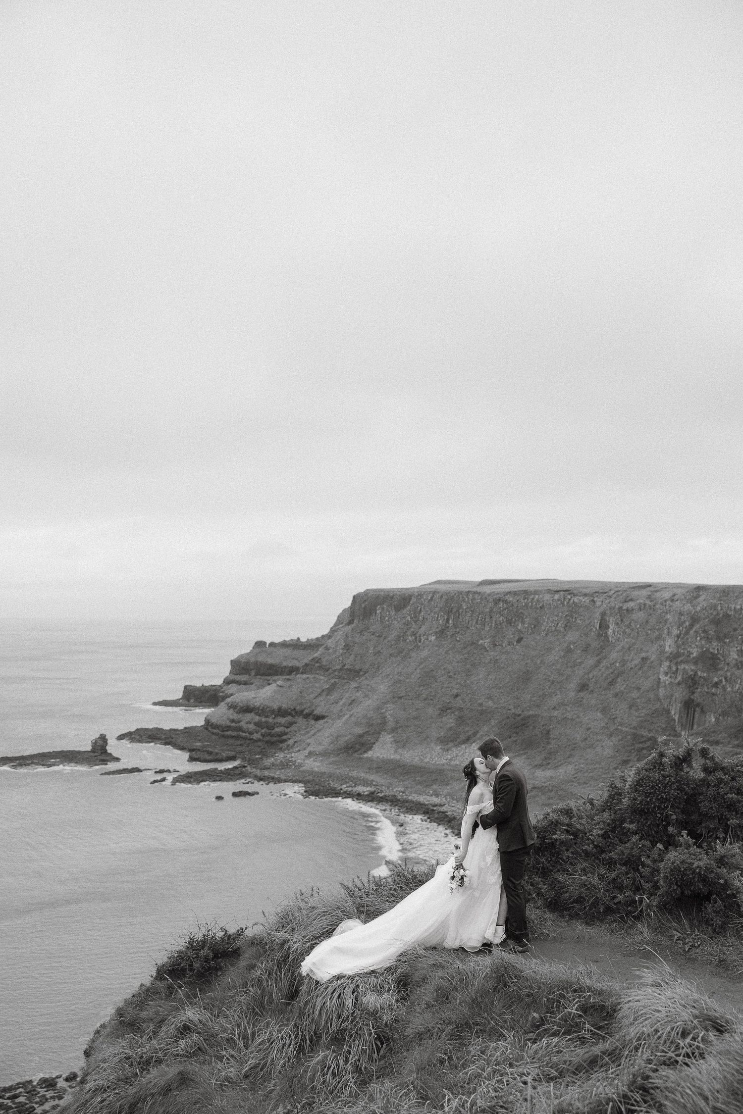 A couple in wedding attire stands at the edge of dramatic coastal cliffs overlooking the ocean on a misty day.