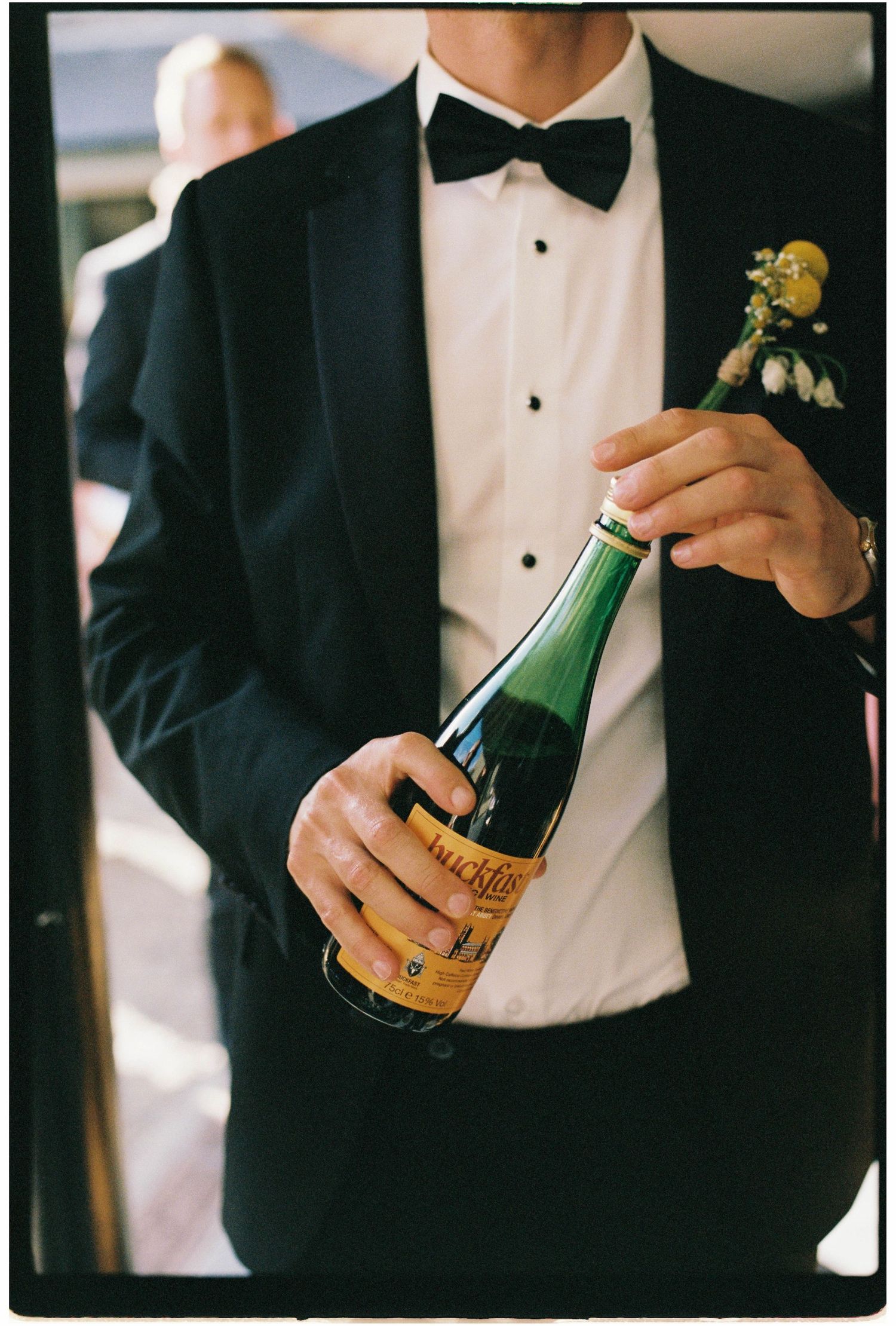 A groom in a black tuxedo with boutonniere holds a bottle of Hucklberry champagne at a wedding celebration.