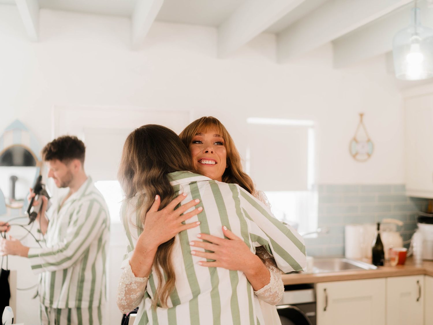 Two friends embracing warmly in a bright modern kitchen wearing matching striped pajamas.