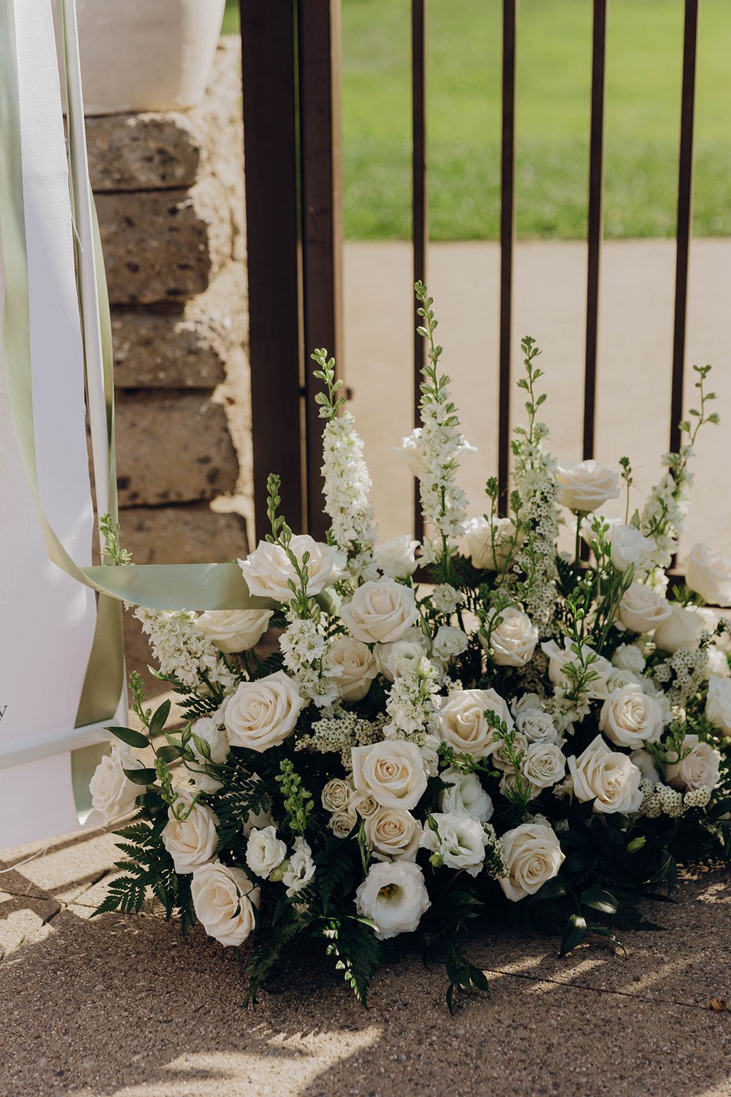 Elegant white rose and delphinium floral arrangement sits by a black iron gate on concrete steps.