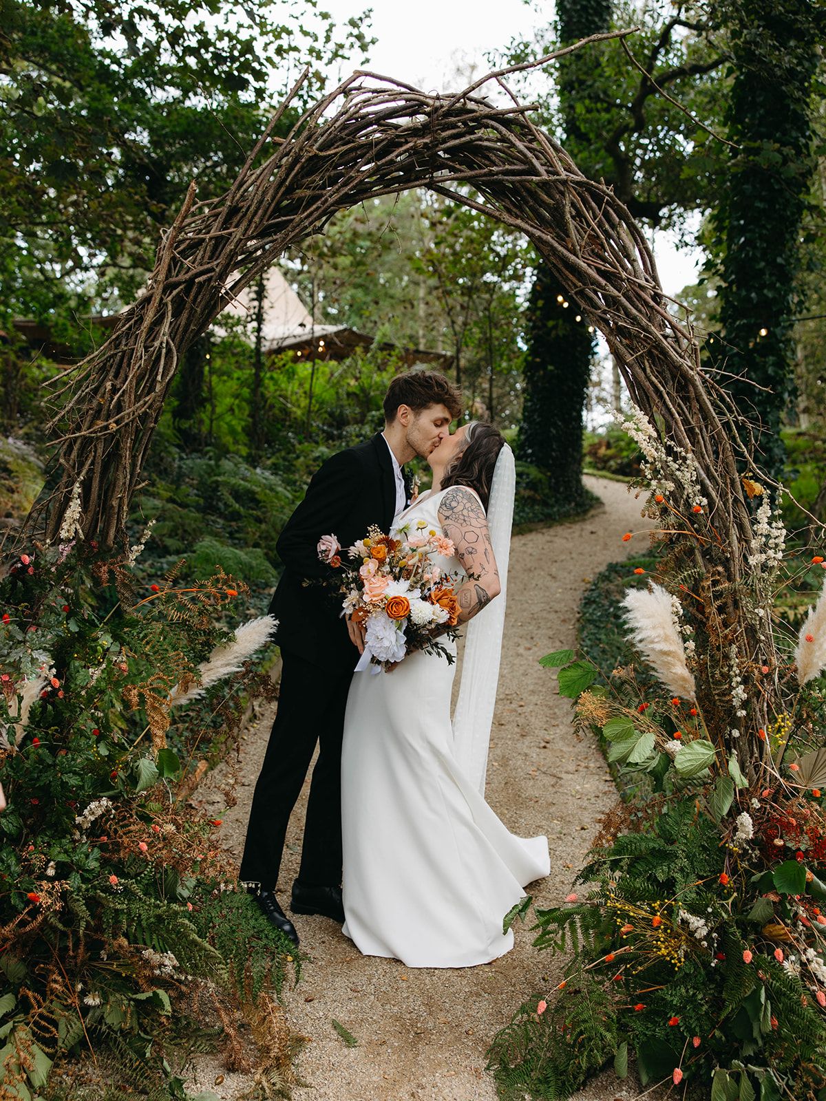 A rustic wedding arch made of twisted branches frames a couple embracing on a garden path surrounded by greenery.