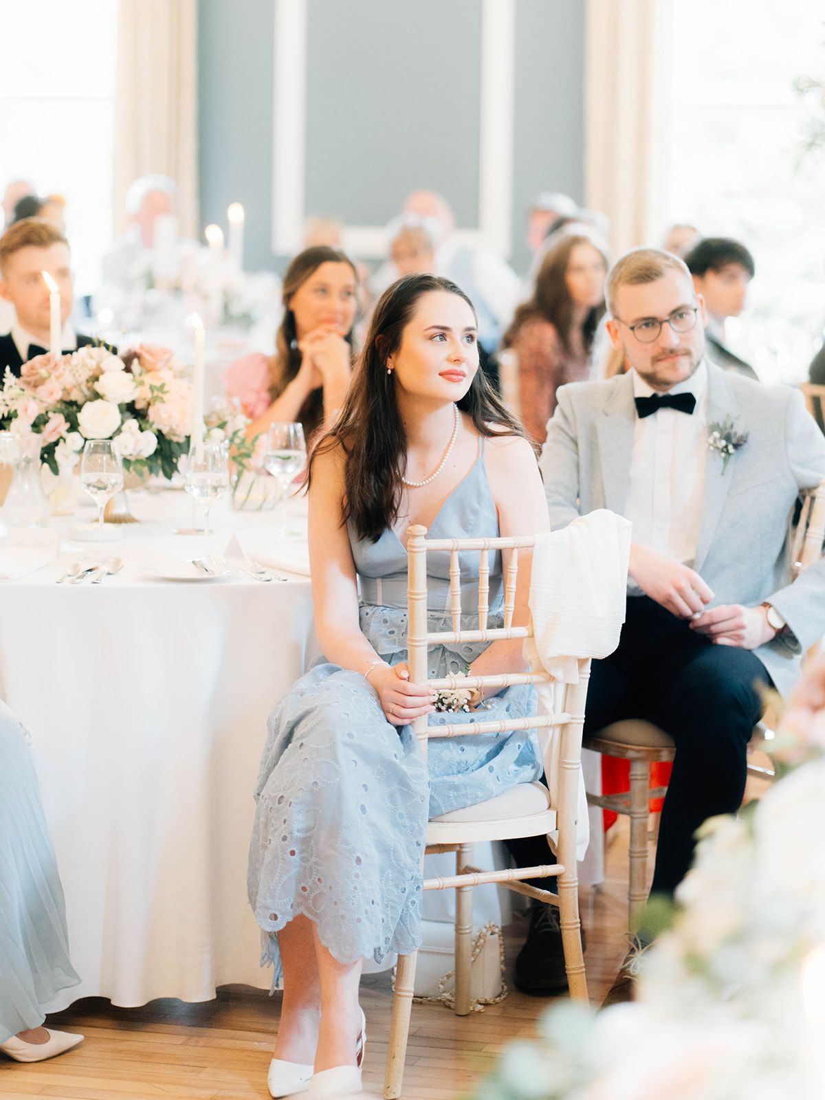 Wedding guests sit at elegant round tables with floral centerpieces in a light-filled reception venue.