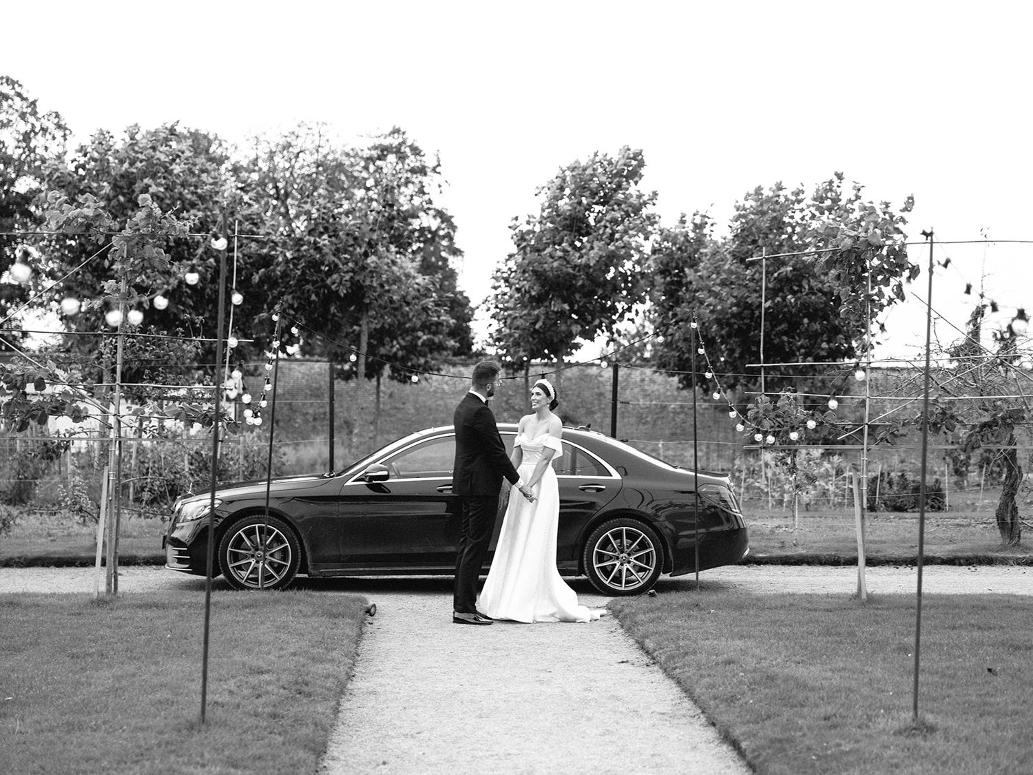Black and white wedding photo showing a couple posing next to a luxury car on a garden pathway.