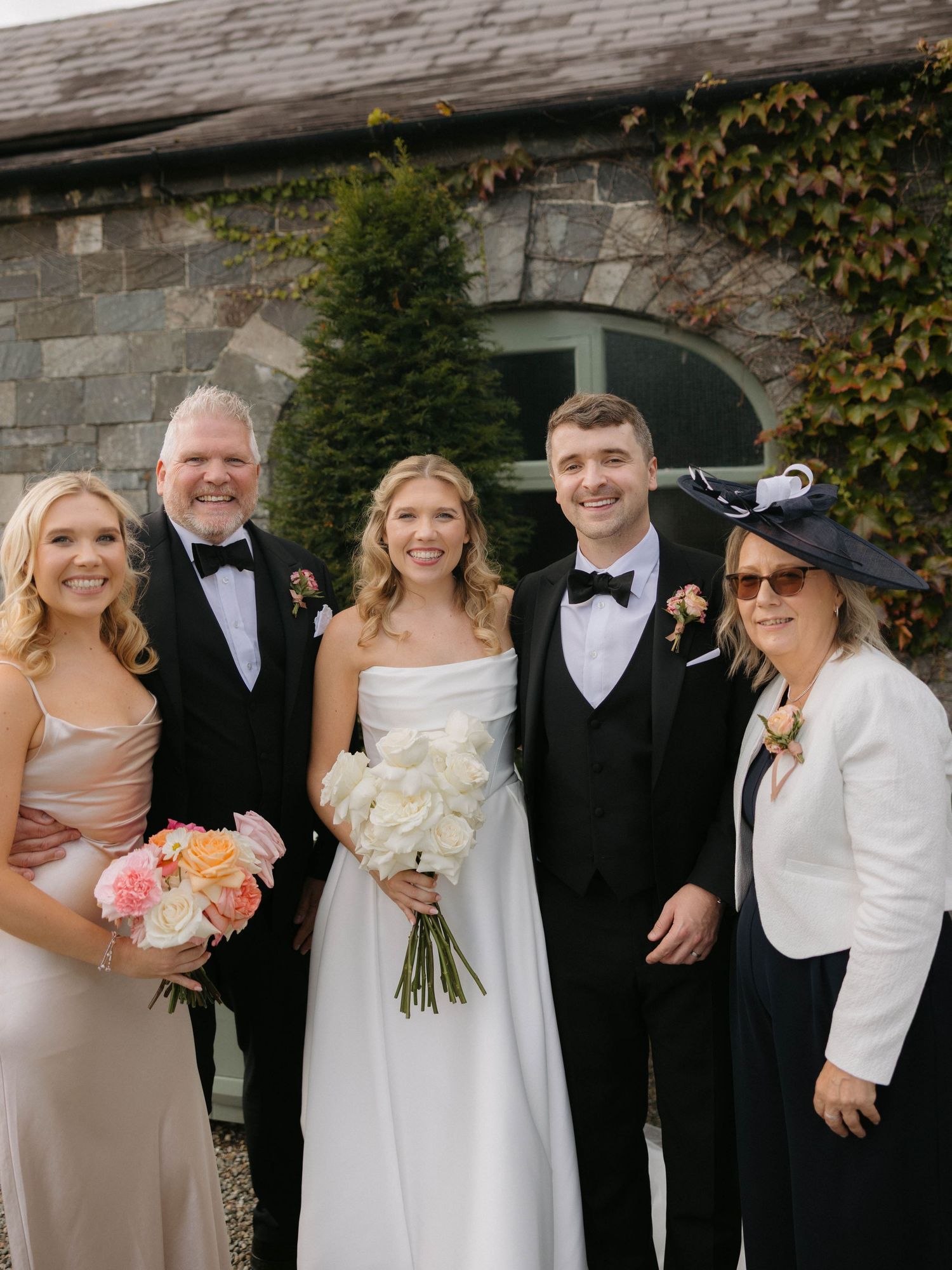 Wedding party group poses outdoors in formal attire with bouquets.