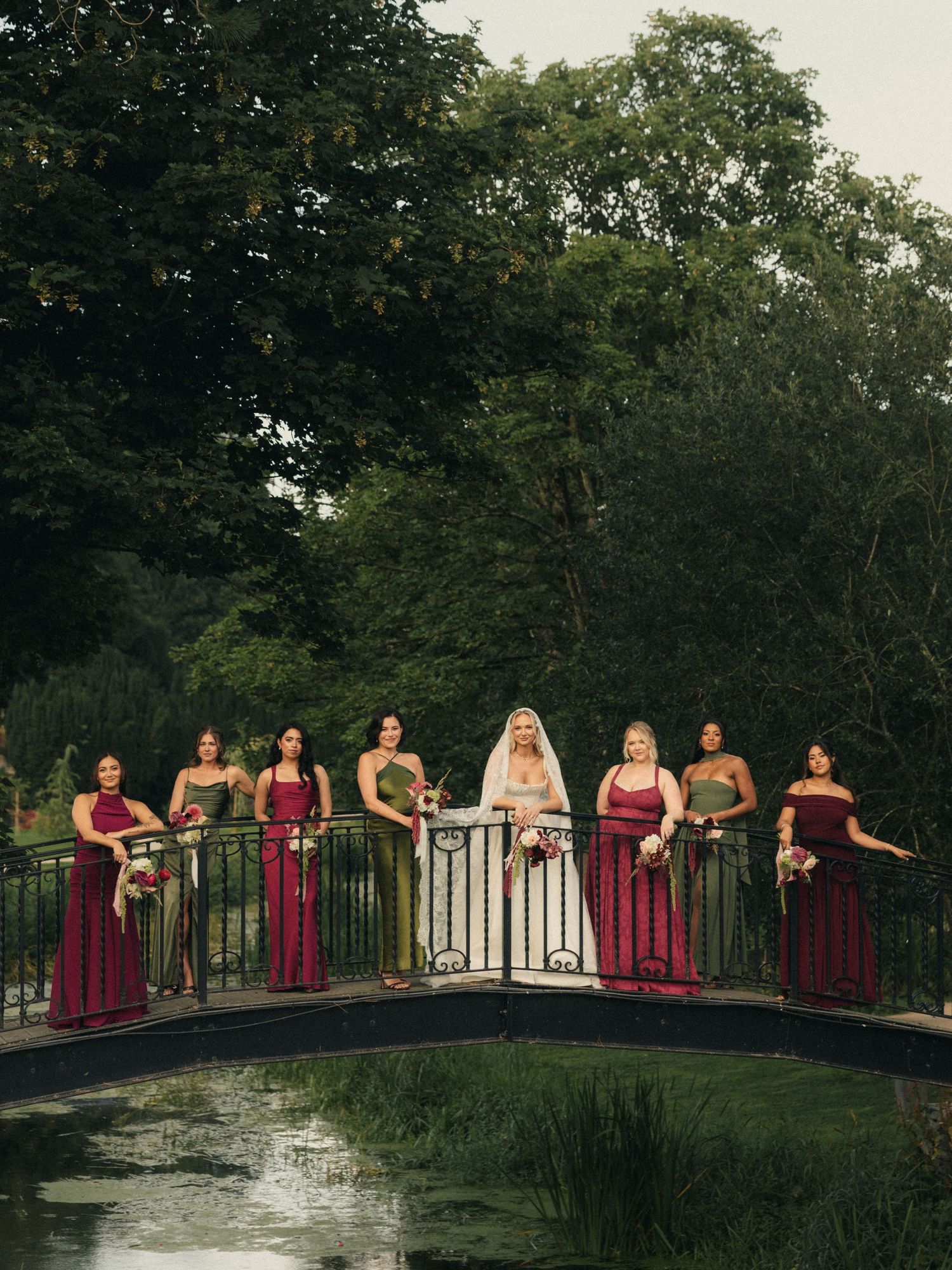 Bride in white gown and veil poses with bridesmaids in burgundy dresses on ornate bridge over pond.