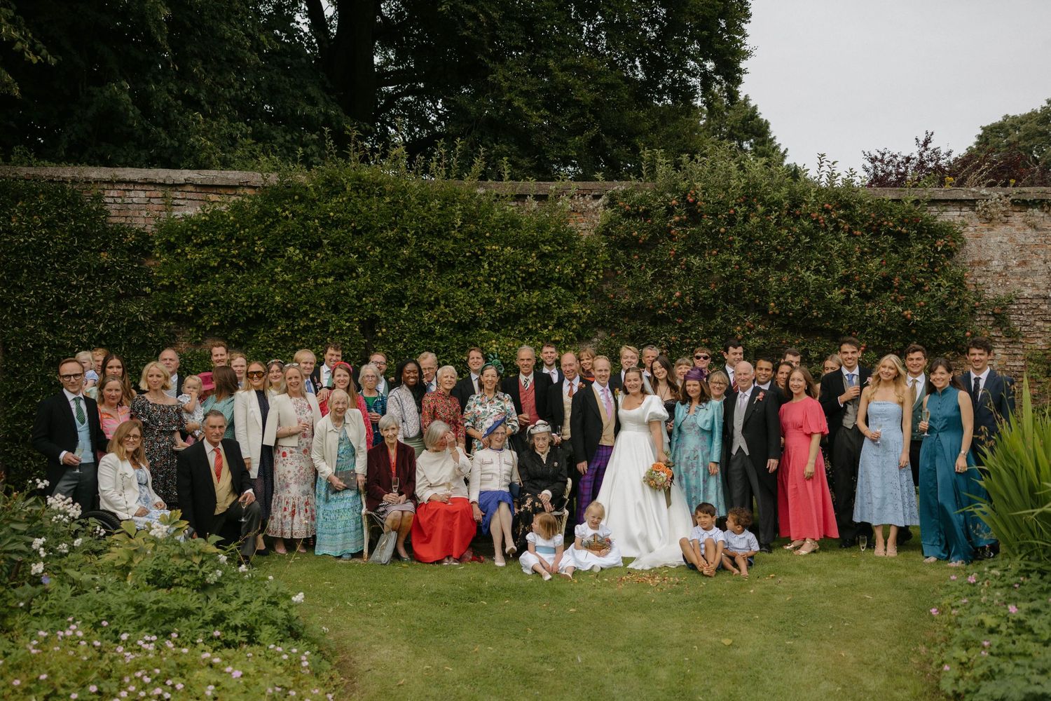 A panoramic collage of wedding guests gathered outdoors in colorful formal attire against a lush green garden backdrop.