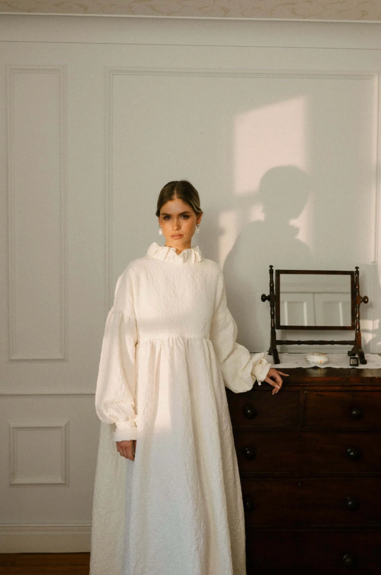 A person in a vintage-style white Victorian dress with puffy sleeves stands next to an antique dresser in a minimalist room.