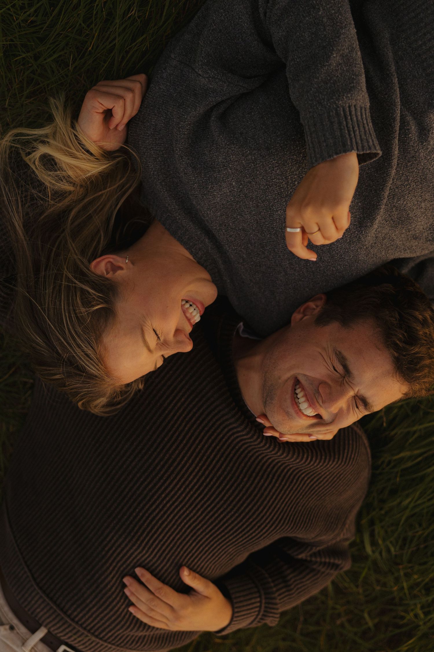 A couple lying on grass sharing joyful laughter in dark sweaters.