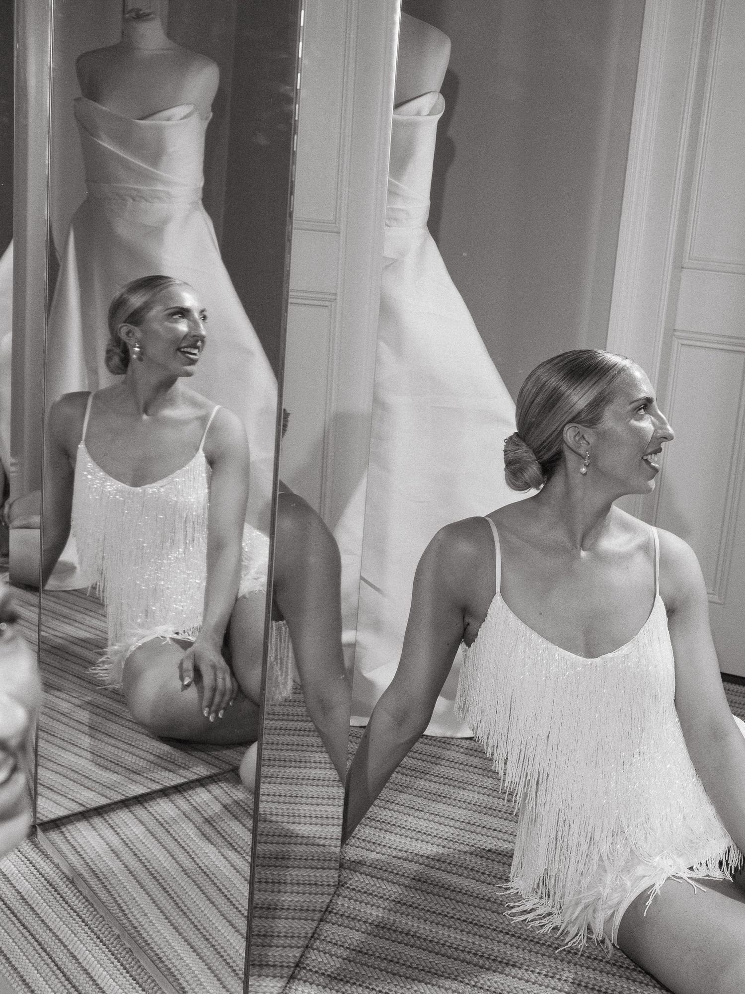 Woman in white fringed top sits on floor before mirror reflecting bridal gown in elegant black and white photo.