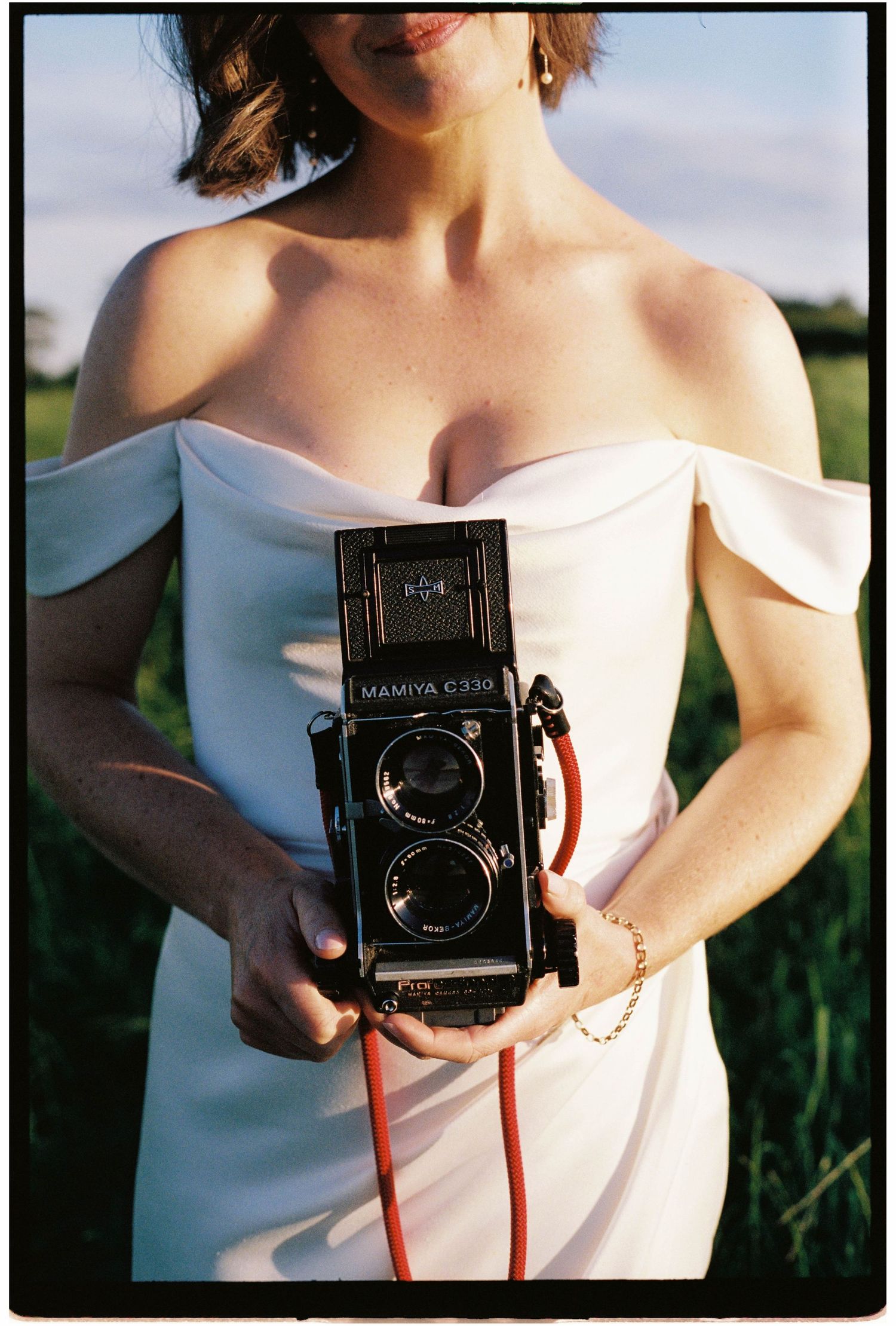 Bride in off-shoulder white wedding dress holding a vintage Mamiya C330 film camera in a green field.