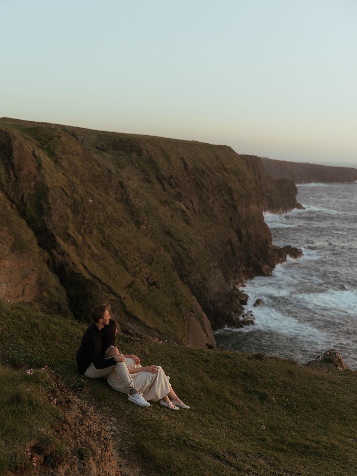 Couple sitting on coastal cliffs overlooking dramatic ocean views at sunset.