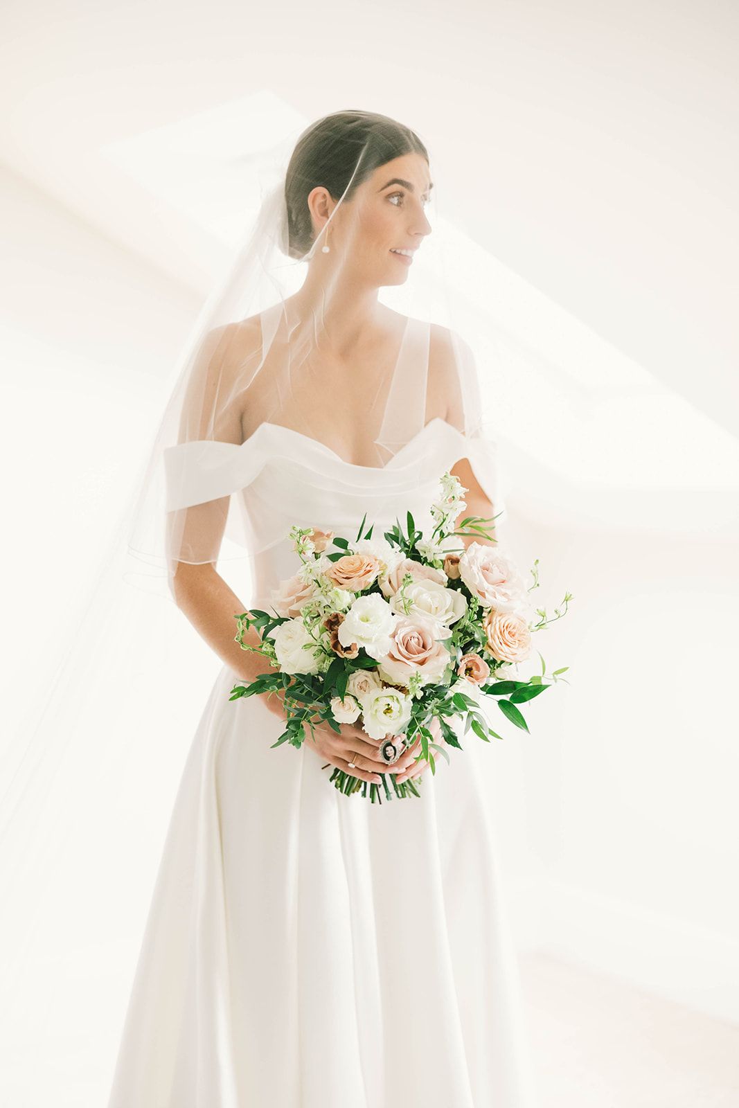 Elegant bride in off-shoulder white wedding gown holds romantic bouquet of blush roses and greenery against bright background.