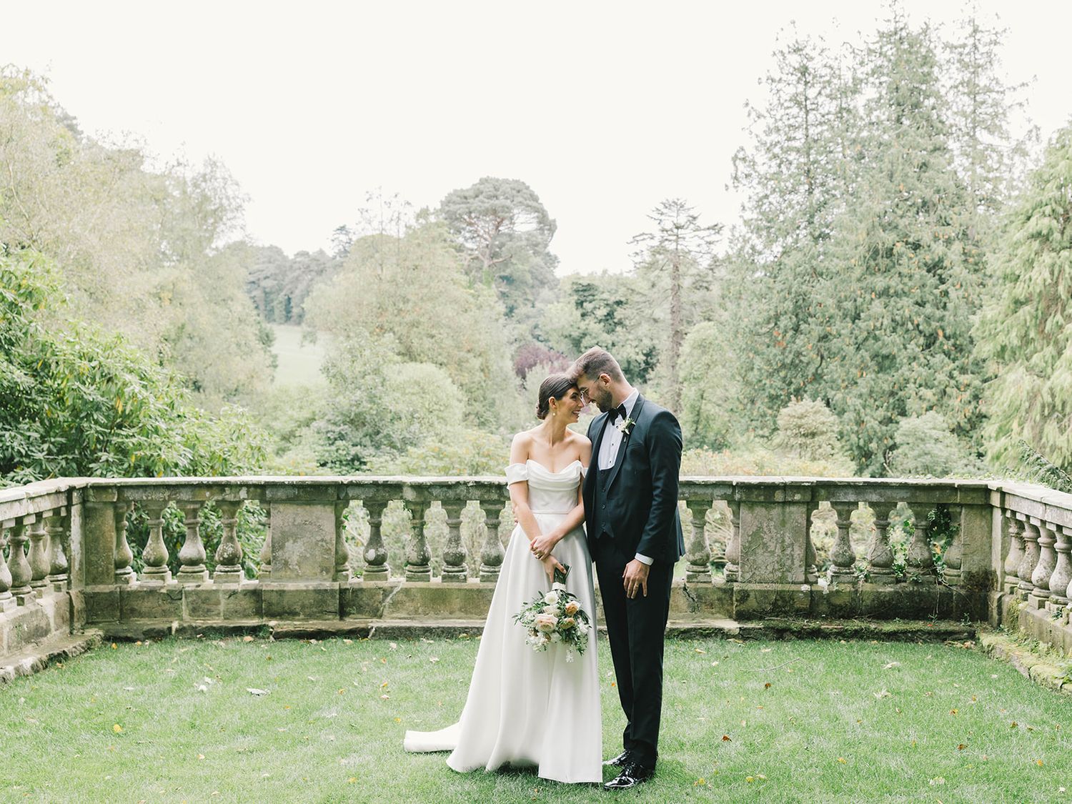 Elegant wedding couple embraces on a stone balcony terrace surrounded by lush garden greenery.