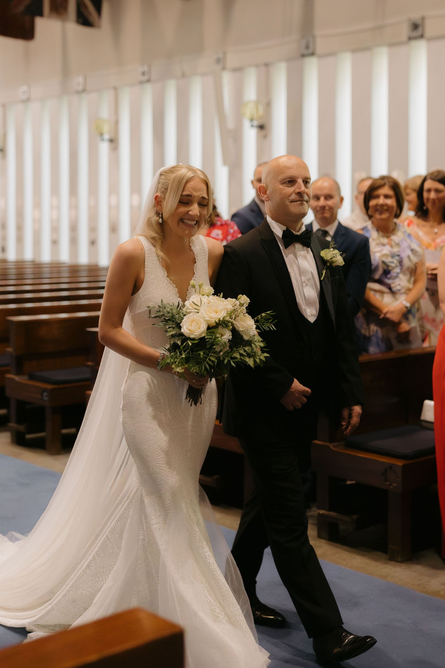 Wedding ceremony moment showing bride and father walking down the church aisle while guests watch from wooden pews.