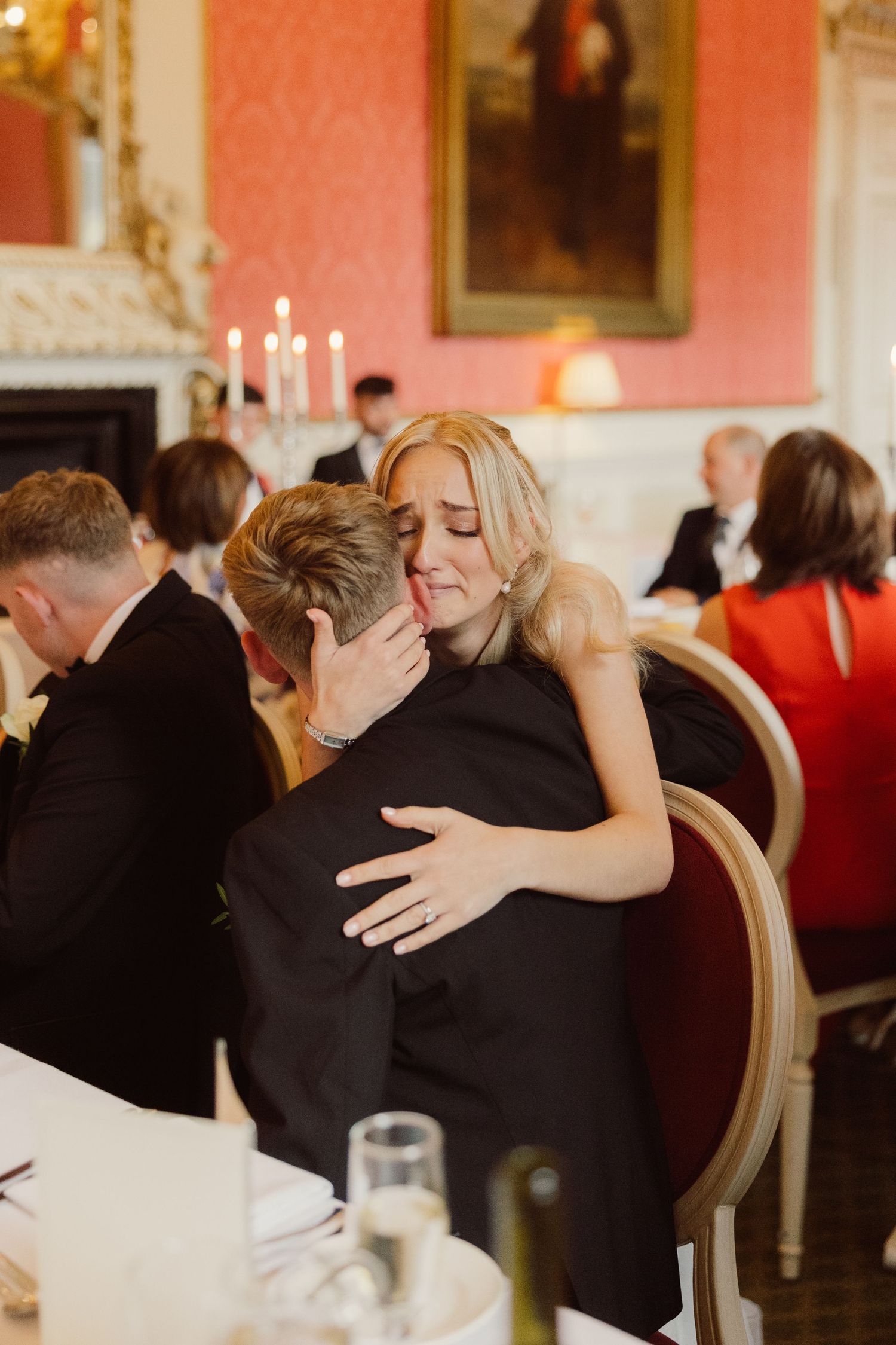 Two guests share an emotional embrace at an elegant dinner party with pink walls and framed artwork in the background.