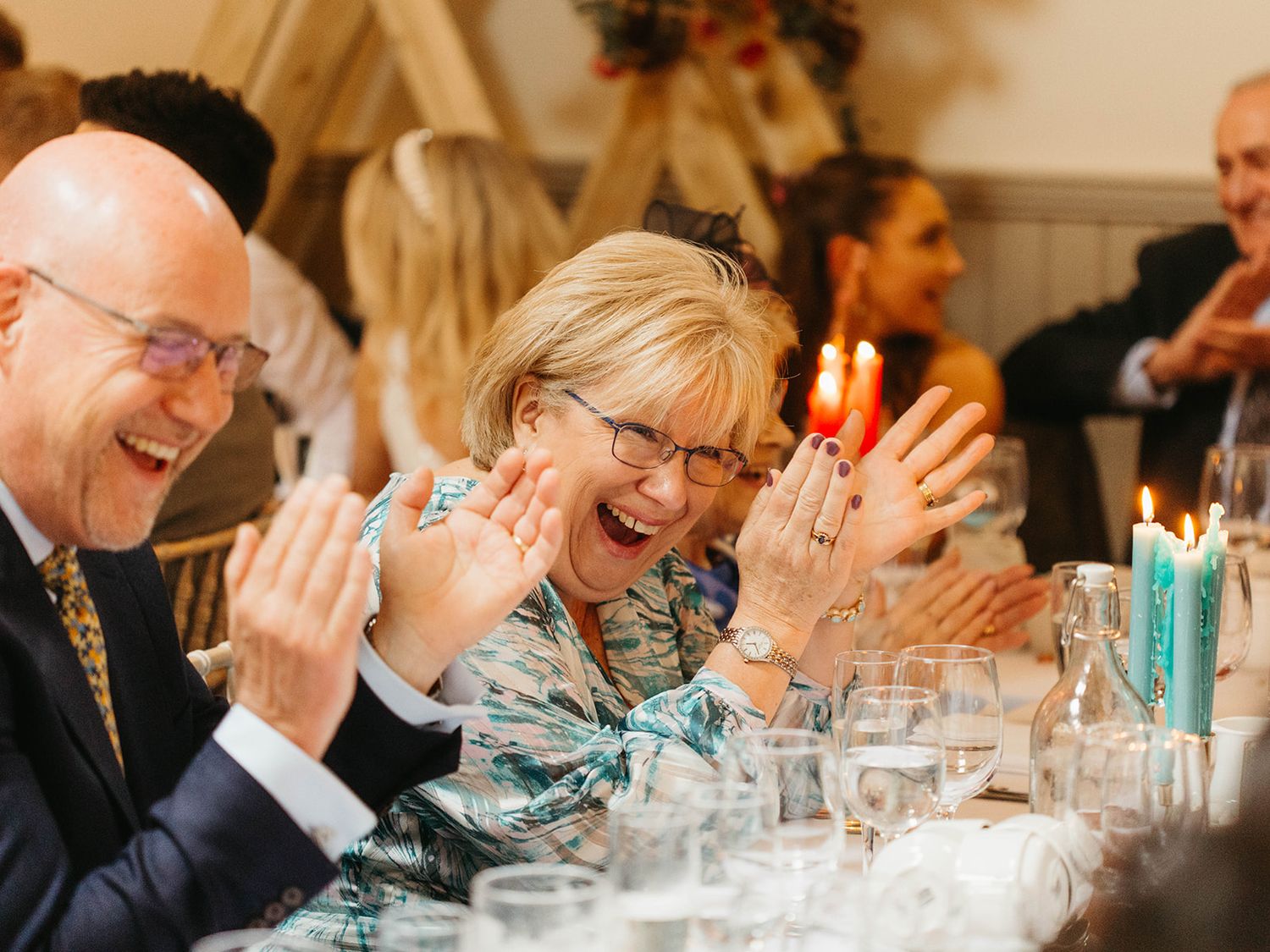 Dinner party guests laugh joyfully while clapping at a formal table setting with candlelight and wine glasses.