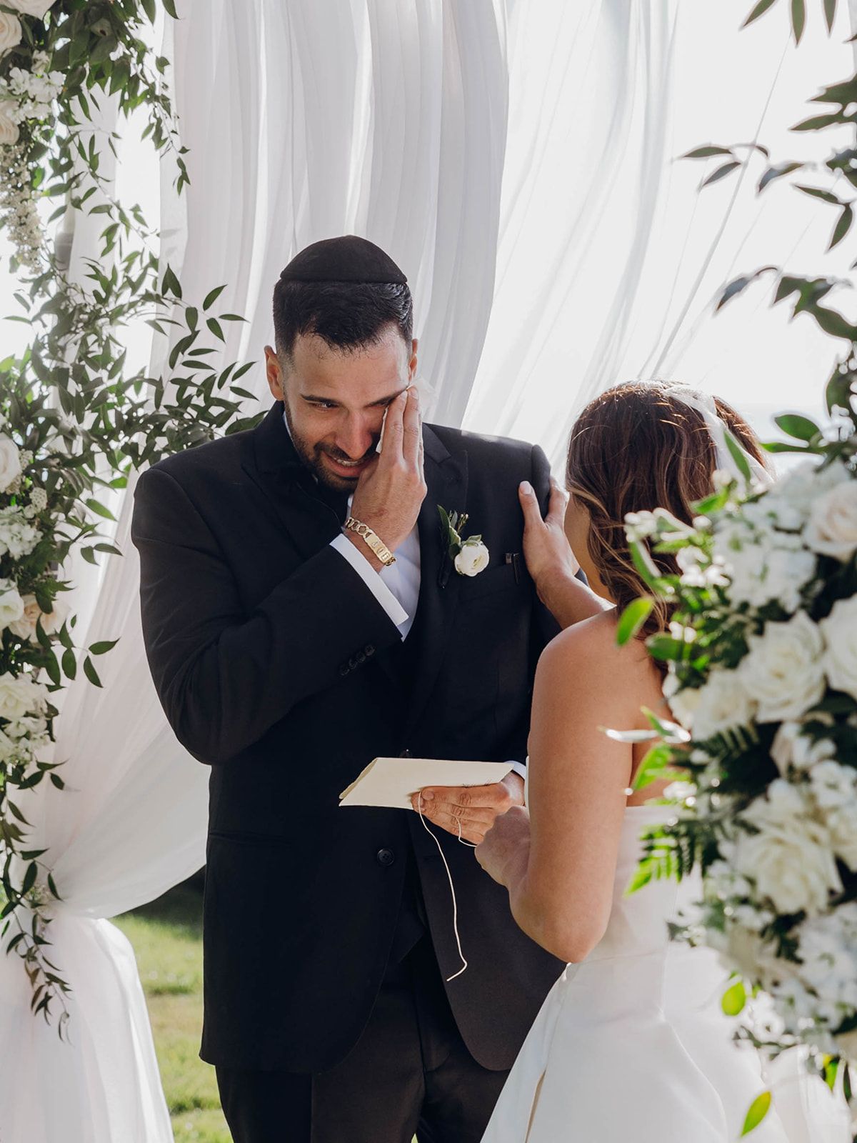 Emotional wedding ceremony moment under a floral arch with white draping and greenery decorations.
