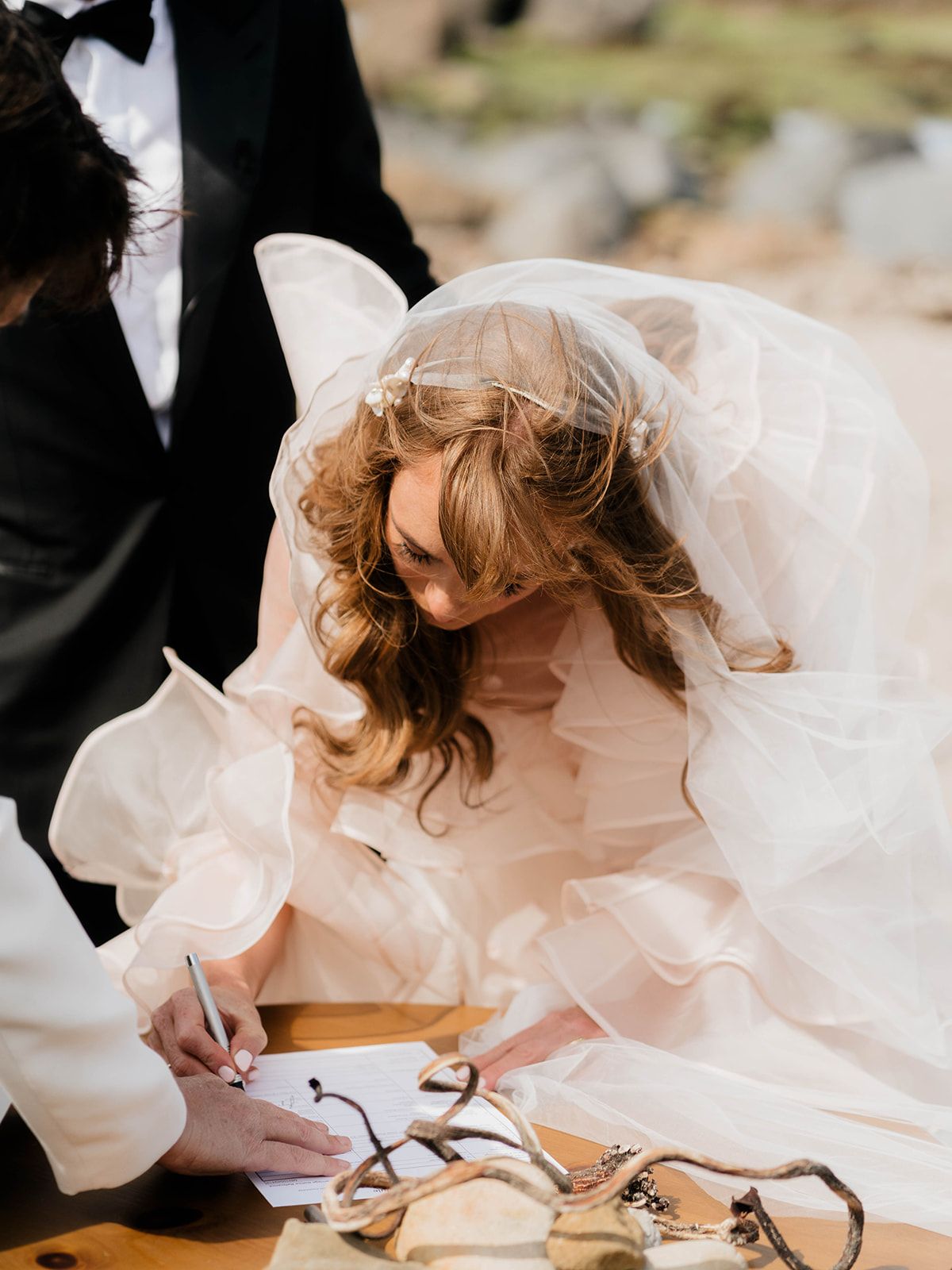 Bride in flowing dress signs wedding documents at outdoor ceremony.