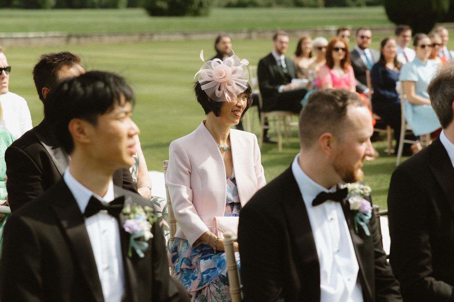 Wedding guests and members of the wedding party in formal attire gather outdoors on a sunny day.