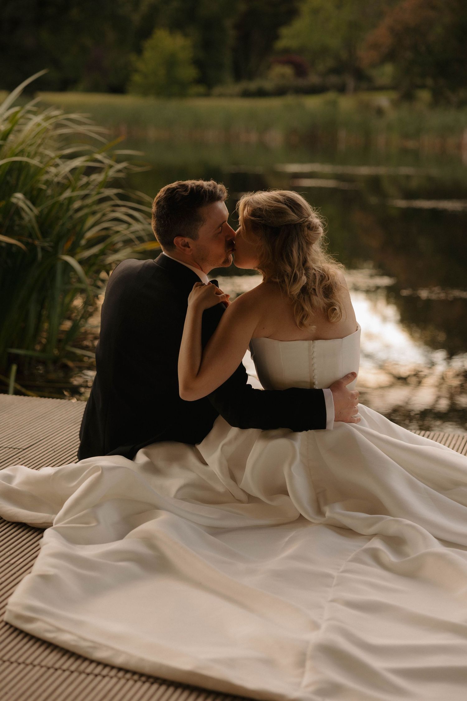 Intimate moment between newlyweds kissing by lakeside at sunset.