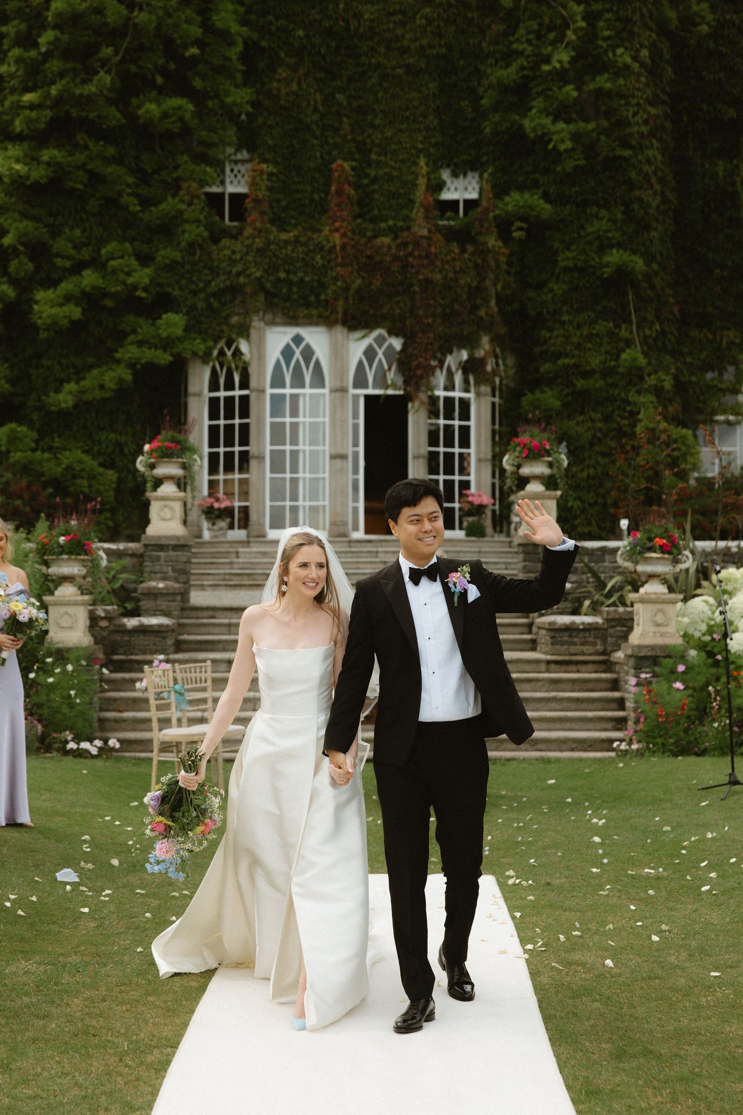 Wedding couple walks hand in hand across a garden lawn with rose petals scattered on the ground at a stone mansion.