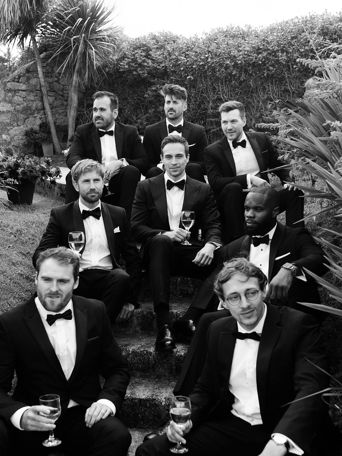 A group of people in formal black tuxedos and bow ties sit casually outdoors among tropical plants.