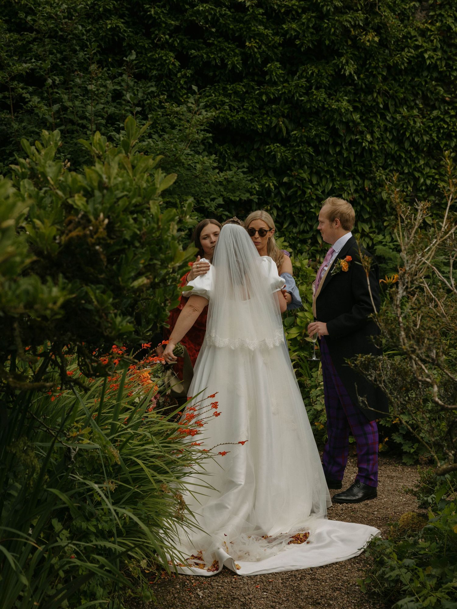 A wedding party gathers in a lush garden path surrounded by greenery and blooming flowers on a sunny day.