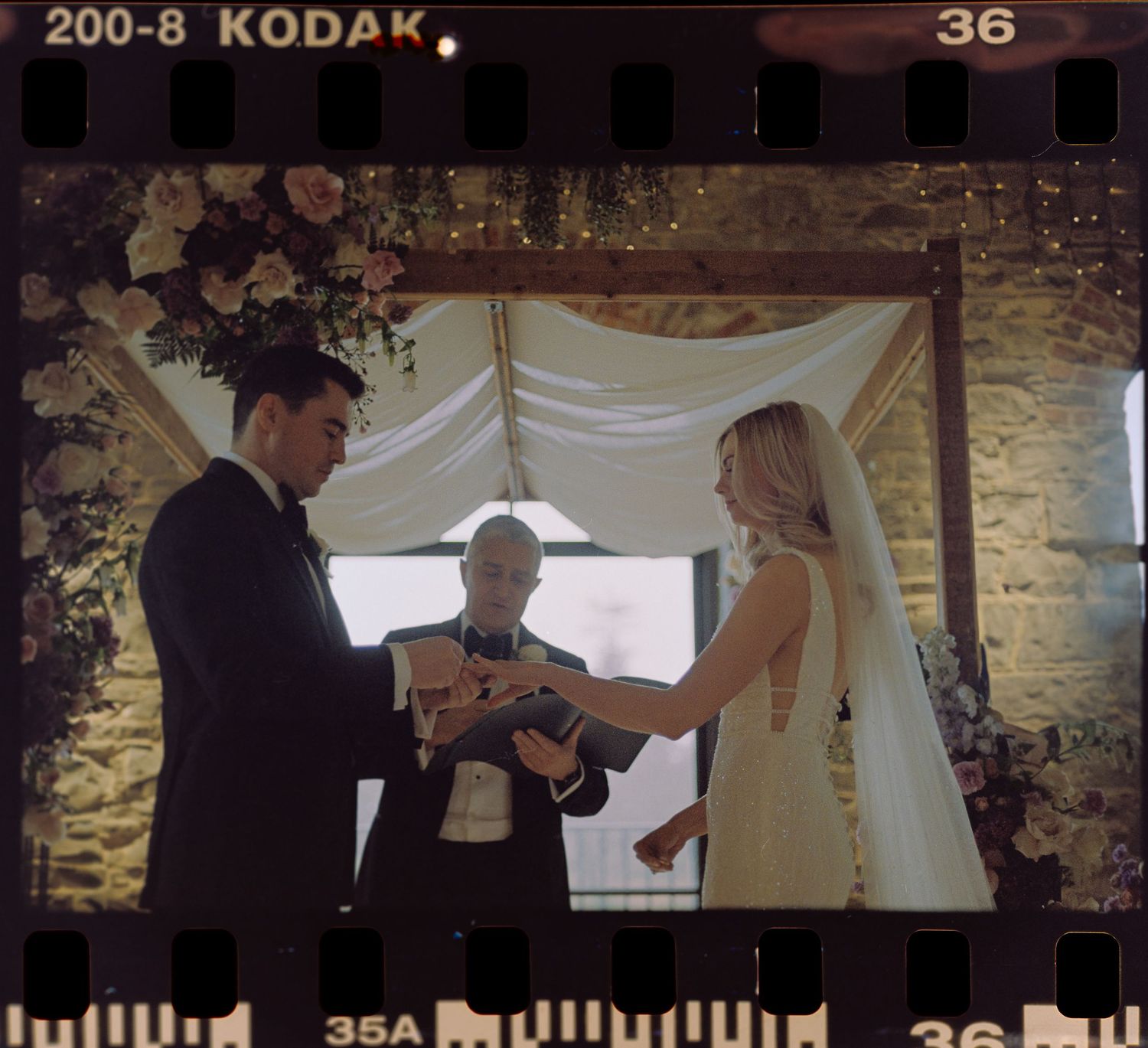 A couple exchanges rings during a wedding ceremony under a white draped canopy with an officiant present.