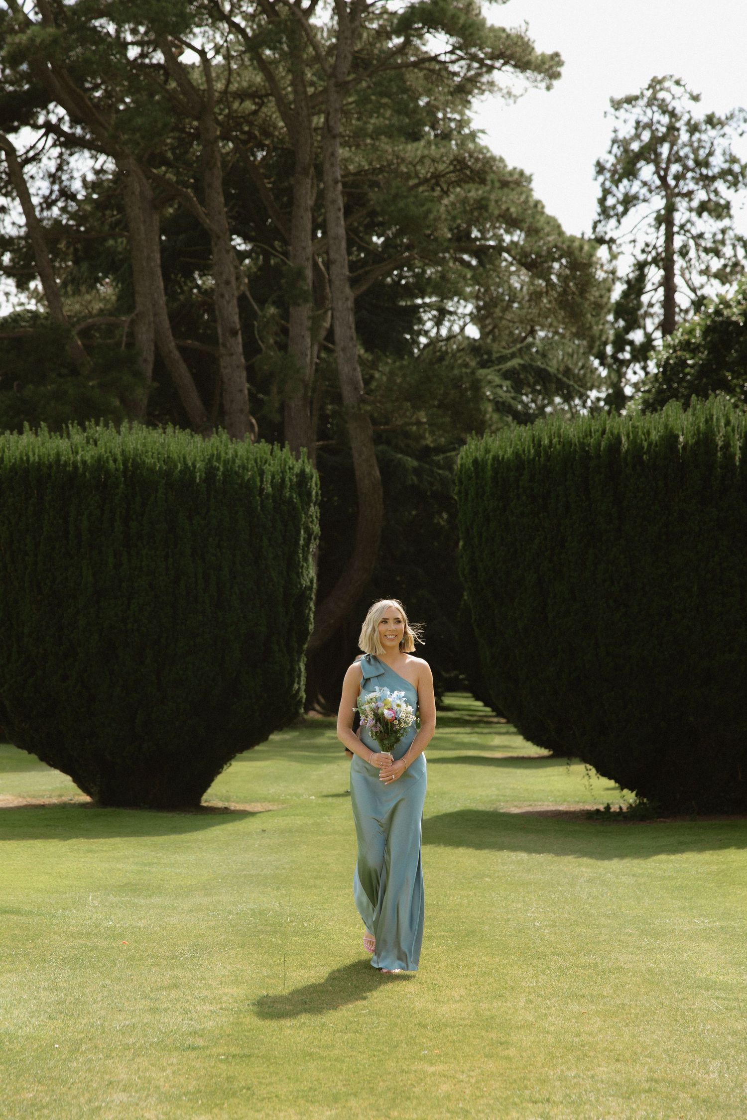 Woman in blue dress walks down garden path between tall hedges on golf course.