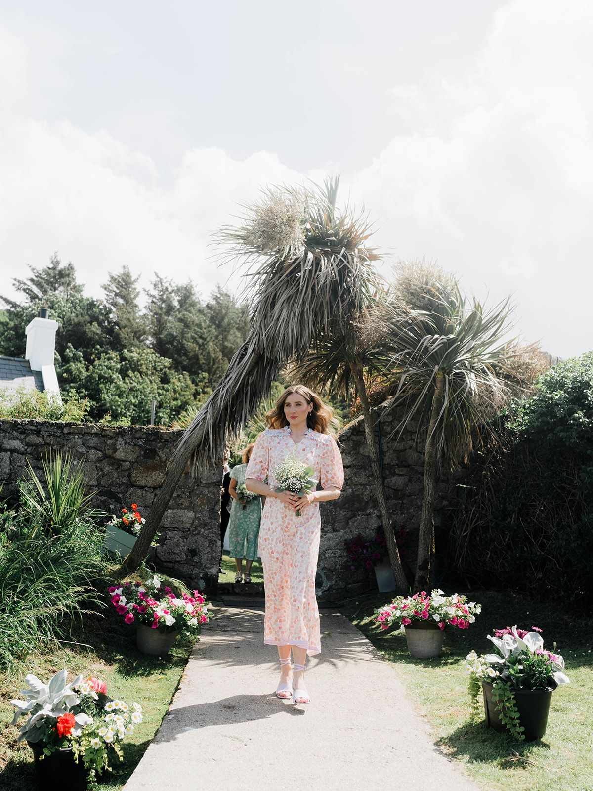 A wedding guest in pink walks along a flower-lined garden path with palm trees.