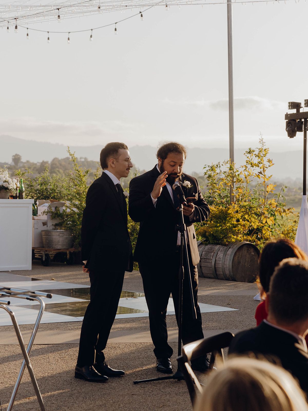 Two men in dark suits standing on an outdoor rooftop venue with string lights and barrels in the background.