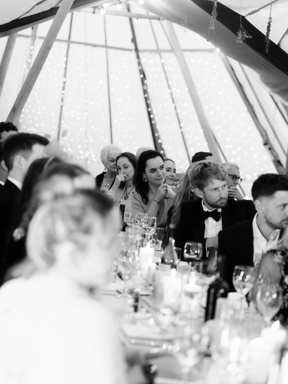 Black and white photo of wedding guests enjoying dinner and speeches inside elegant tent reception.