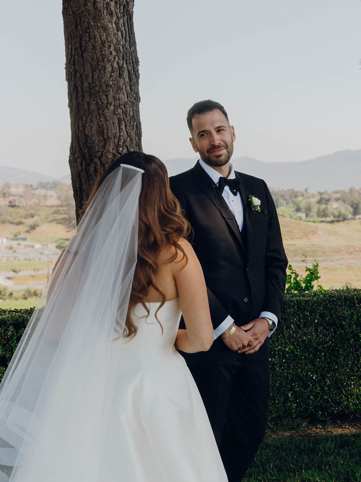 A bride in a white strapless gown and veil stands with someone in a black tuxedo during an outdoor wedding ceremony.