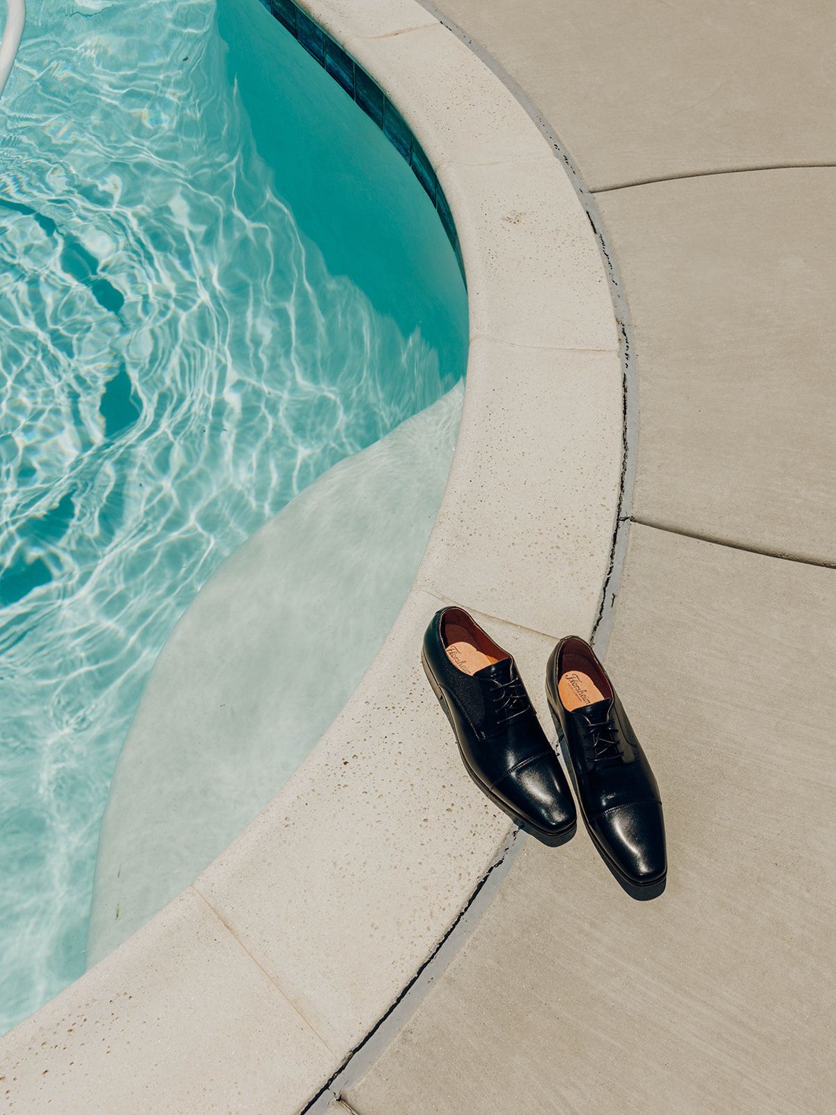 A pair of black leather dress shoes placed on a curved concrete pool edge next to turquoise swimming pool water.