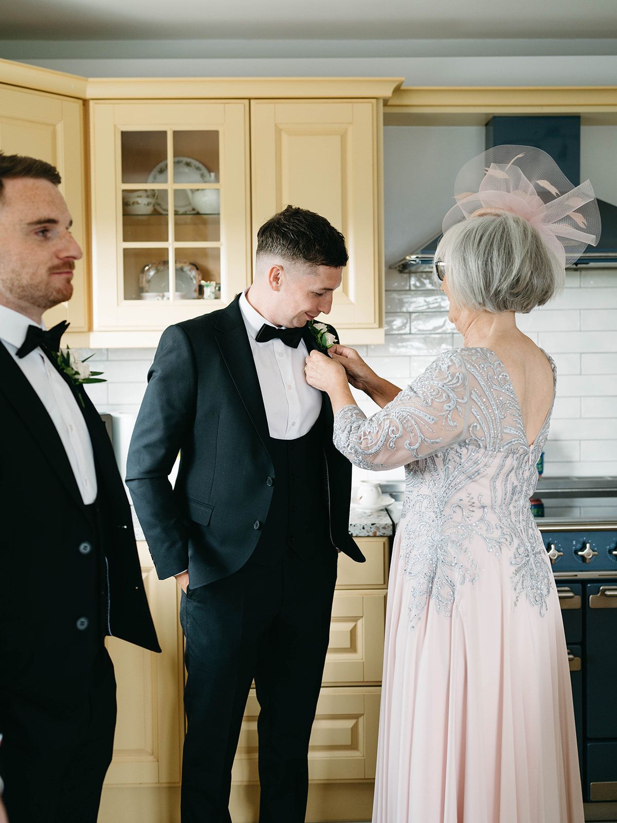 Wedding preparations take place in an elegant kitchen with cream cabinets as formal attire is adjusted.