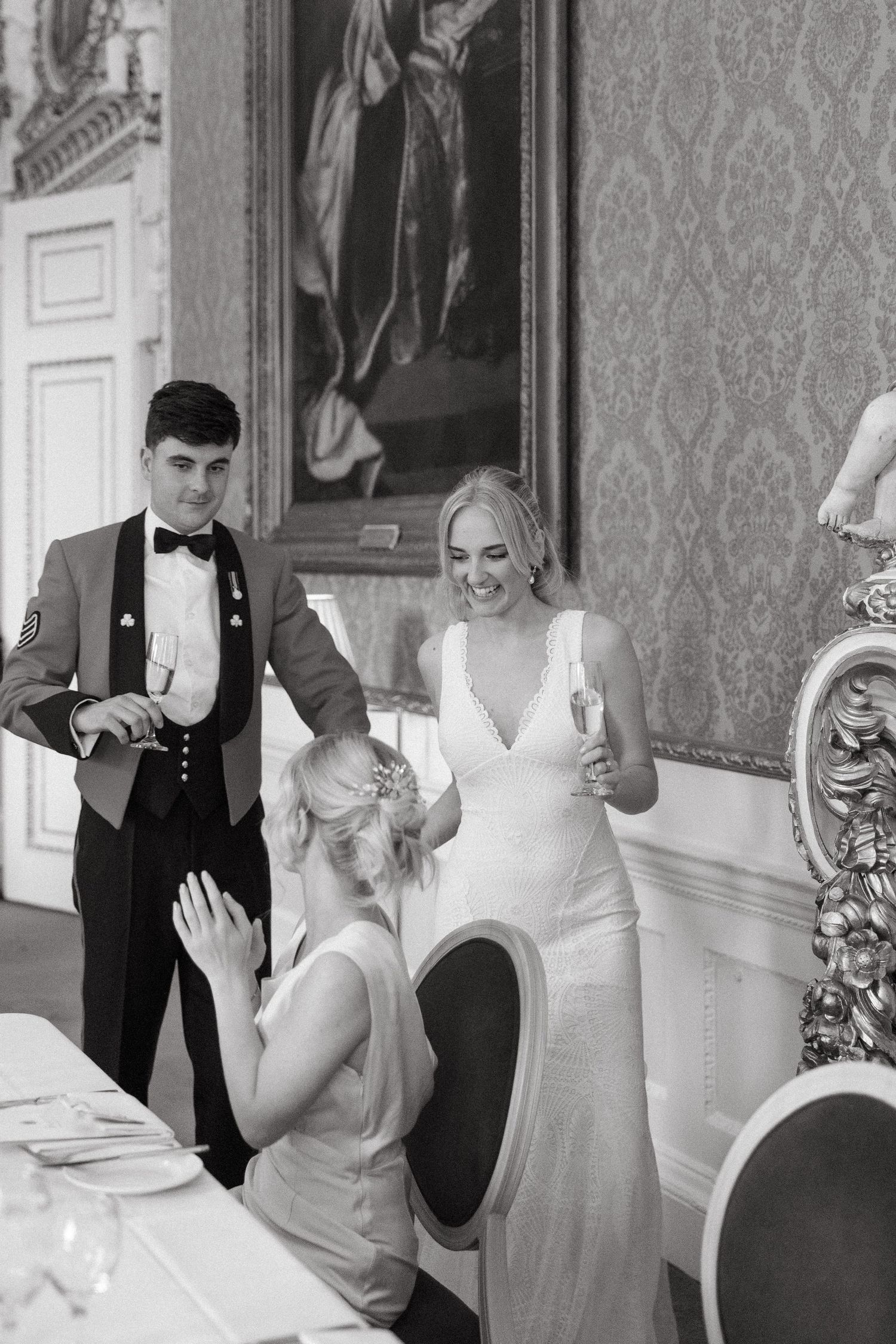 Black and white photo of wedding party guests in formal attire laughing and drinking at an elegant venue.