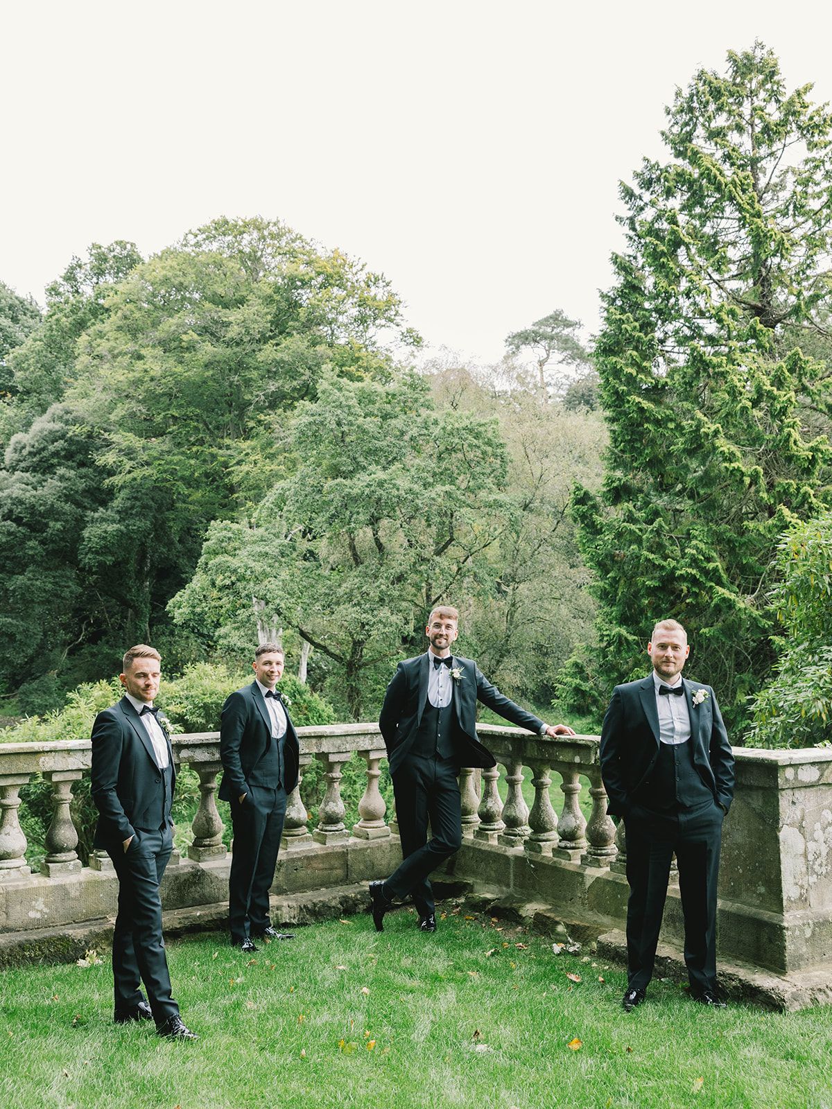 Four people in formal tuxedos pose on a stone balcony with lush green trees in the background.