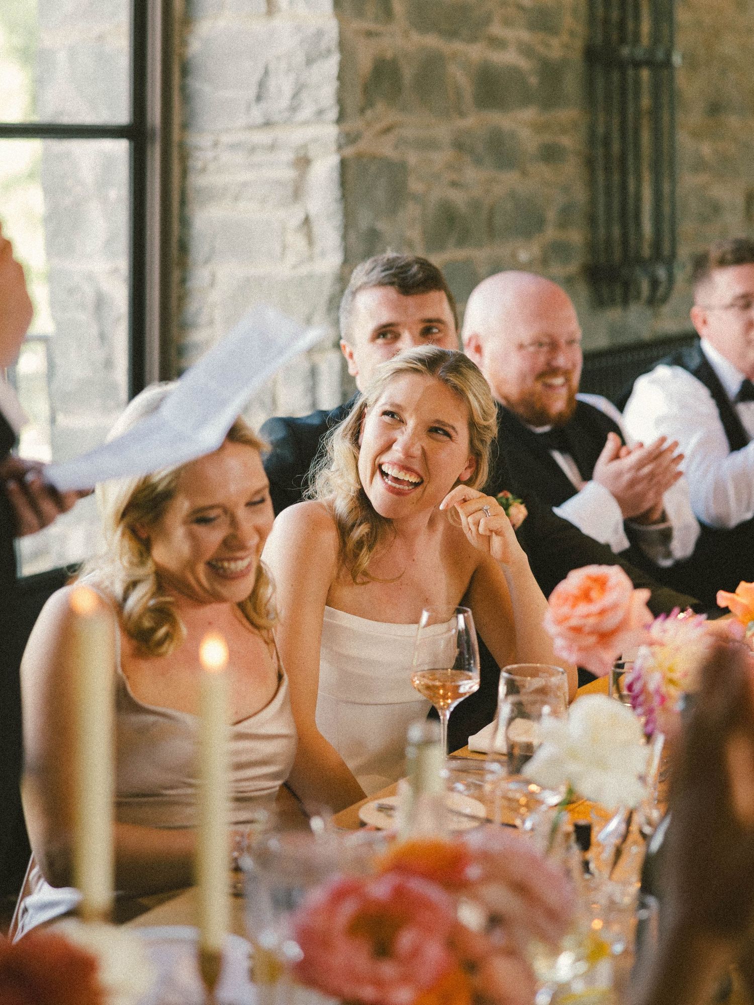 Wedding reception guests laugh and smile during toasts at an elegant stone venue.