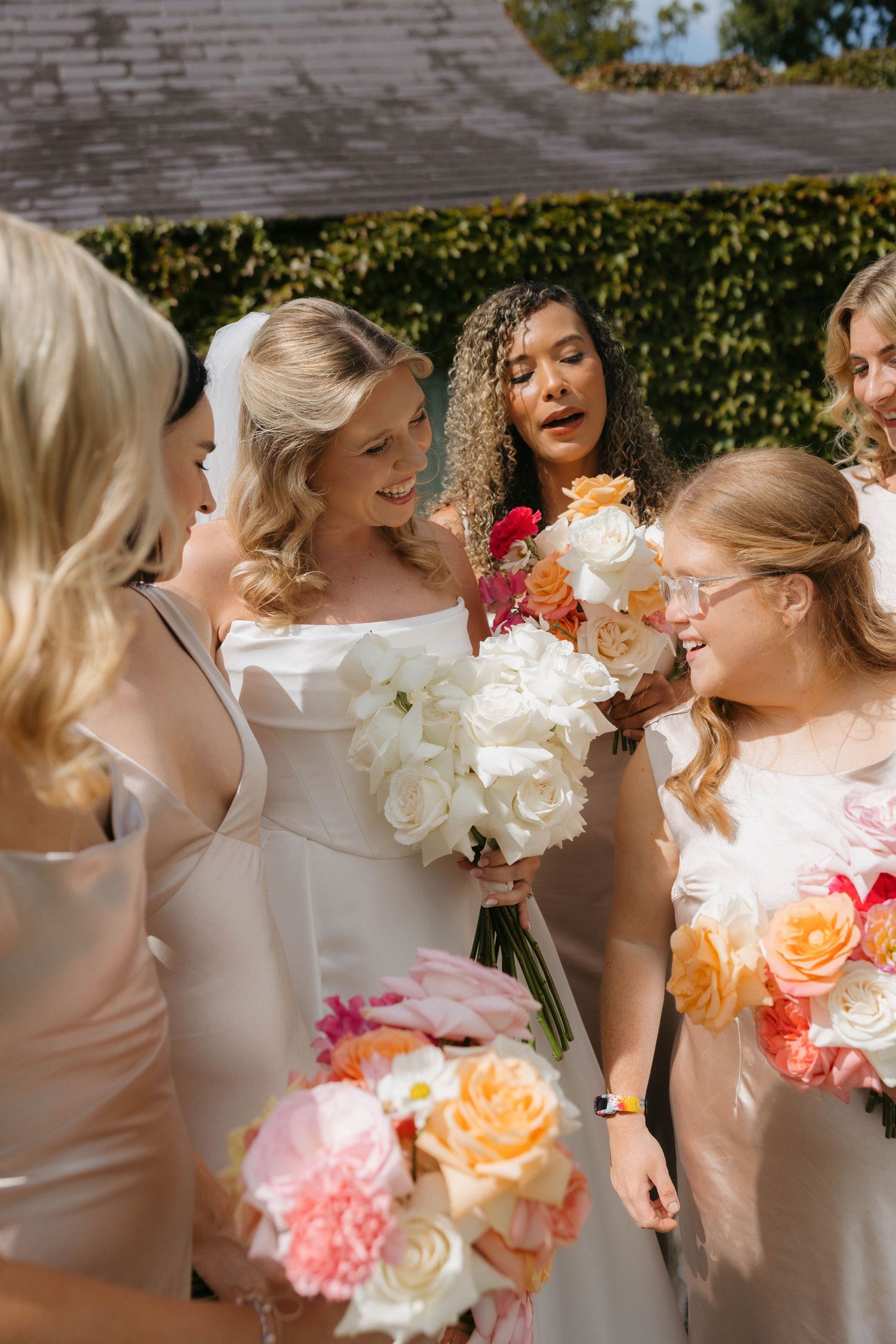 Bridesmaids in white dresses holding vibrant floral bouquets in natural sunlight.