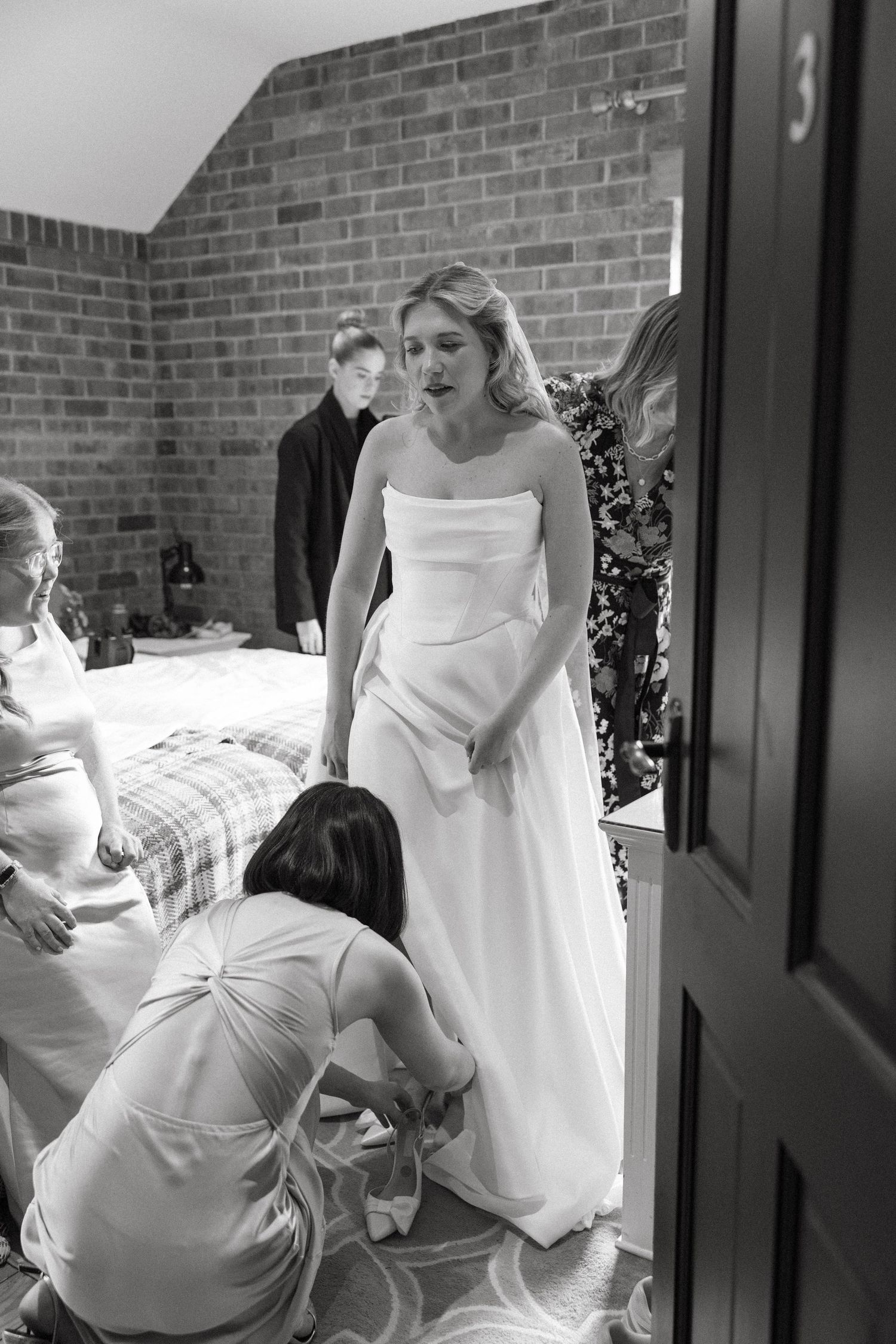Black and white image of wedding dress preparations with helpers assisting the bride.