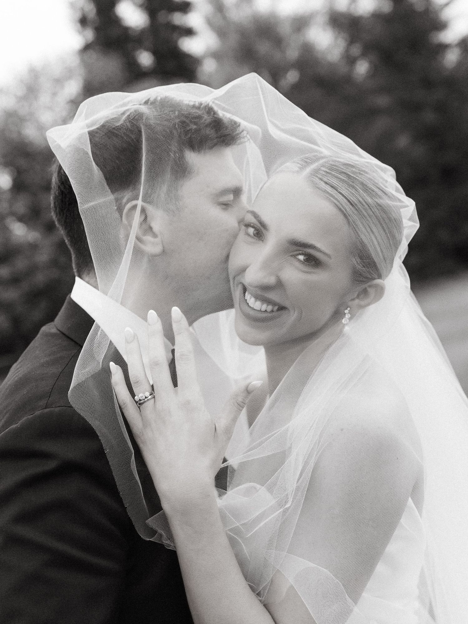 Black and white wedding photo of smiling bride and groom sharing a kiss under her flowing veil.
