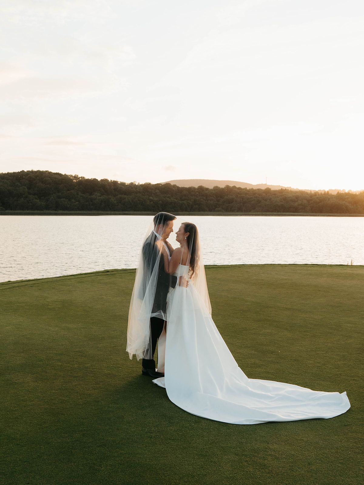 A romantic wedding couple stands on a lush green lawn against a golden sunset backdrop with a flowing white veil trailing behind.