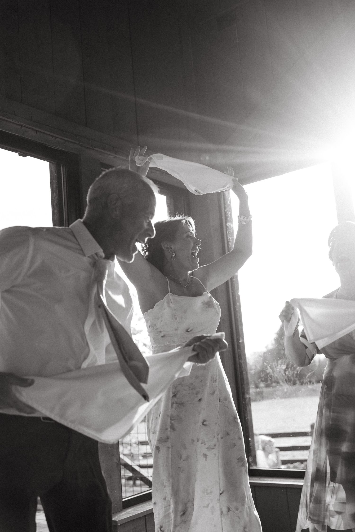 Black and white photo of a couple dancing joyfully at their wedding reception with sunlight streaming through windows.
