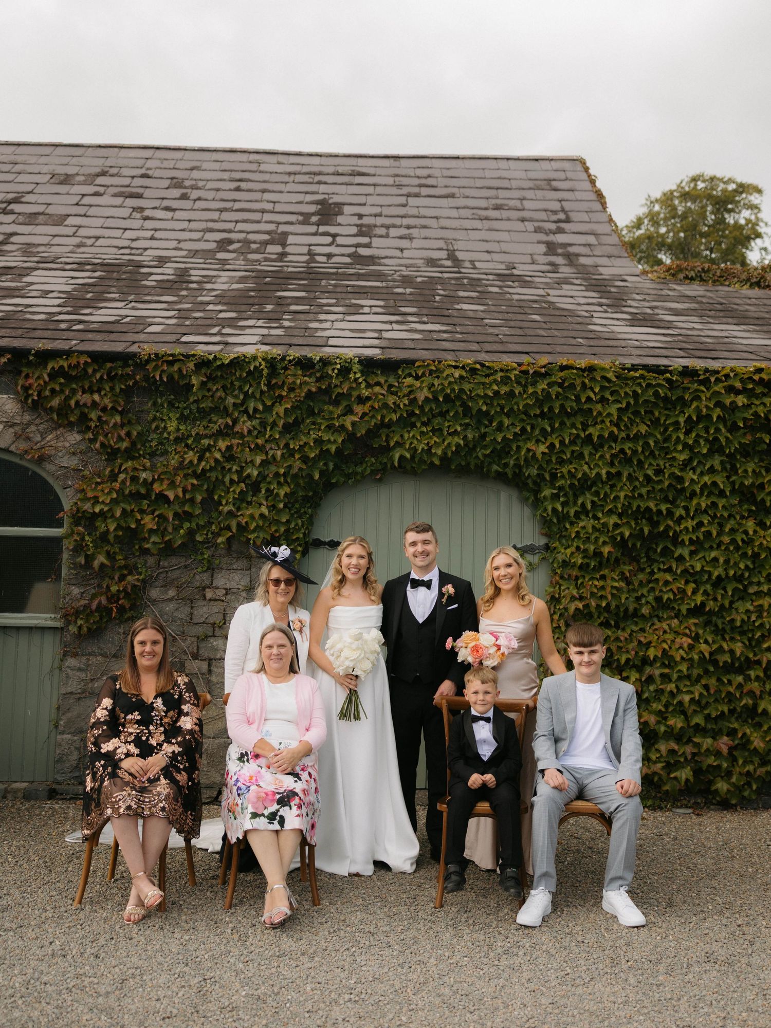 Wedding party members sit casually along an ivy-covered stone wall.